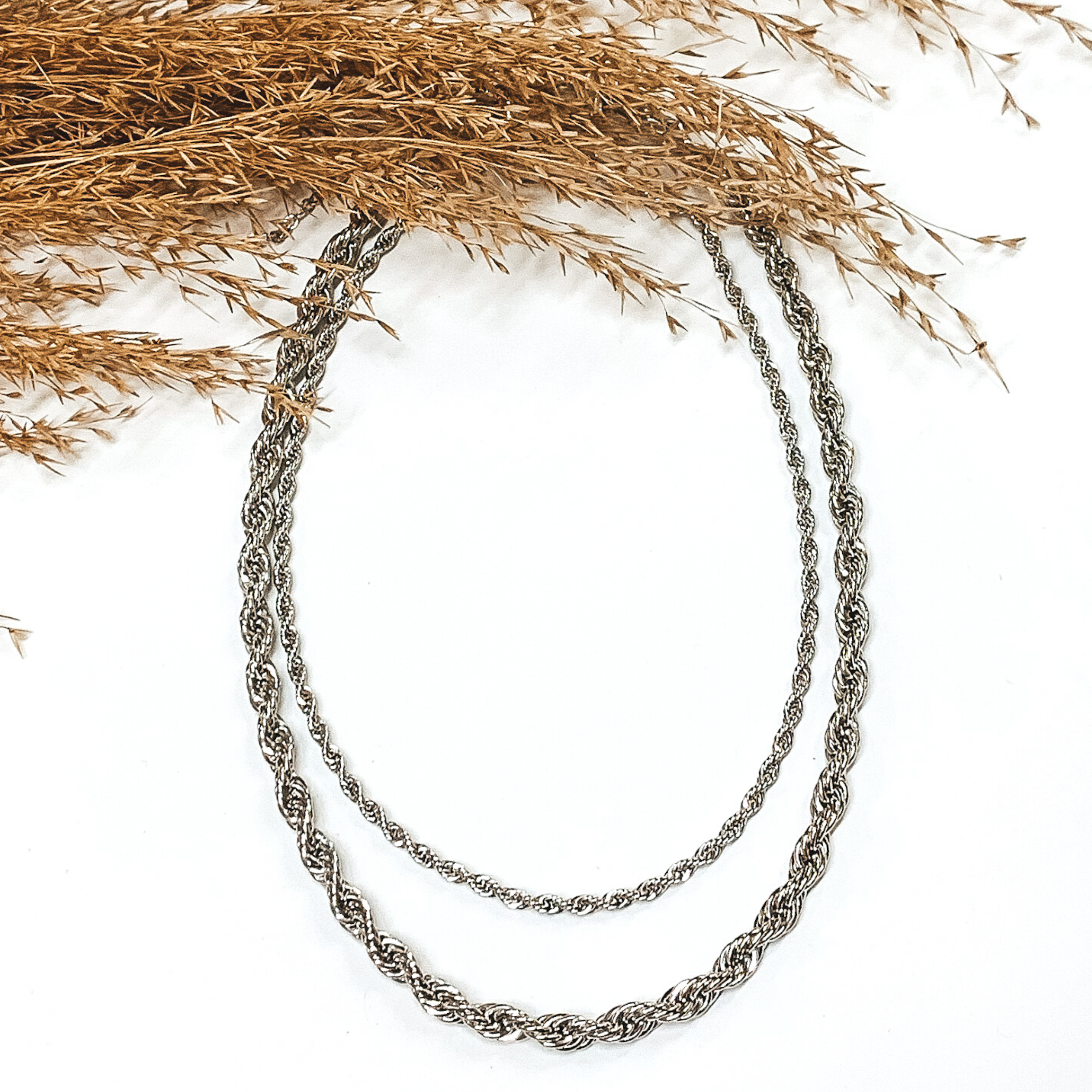 Double layered silver rope chain necklace. This necklace is pictured on a white background with tan floral at the top of the picture.