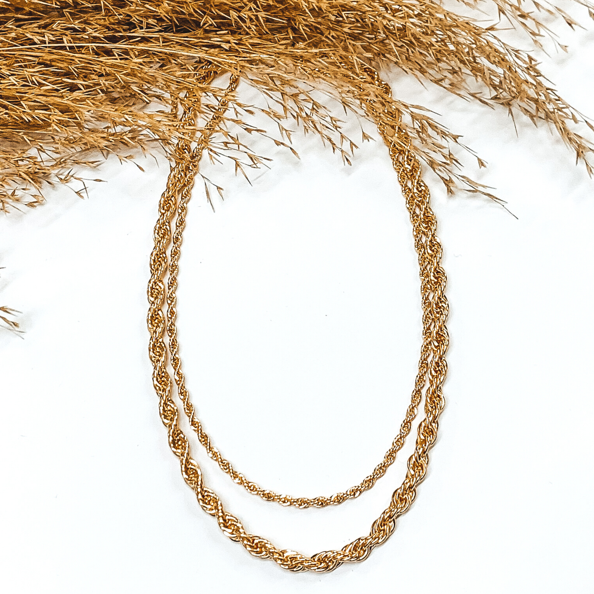 Double layered gold rope chain necklace. This necklace is pictured on a white background with tan floral at the top of the picture. 