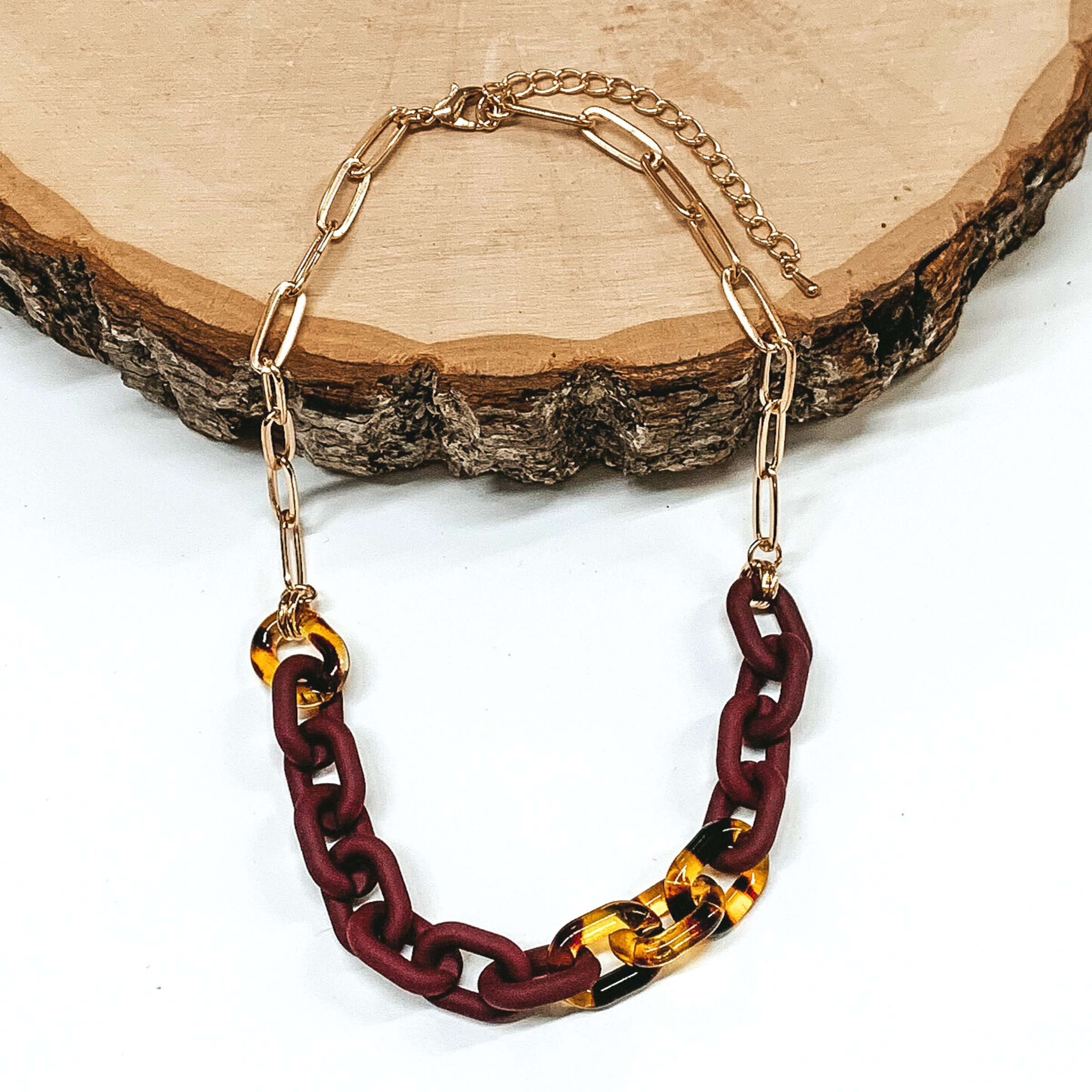 Half gold paper clip chain necklace. The other half is a thick matte maroon chain with a few links on tortoise shell links. This necklace is pictured partially laying on a piece of wood on a white background. 