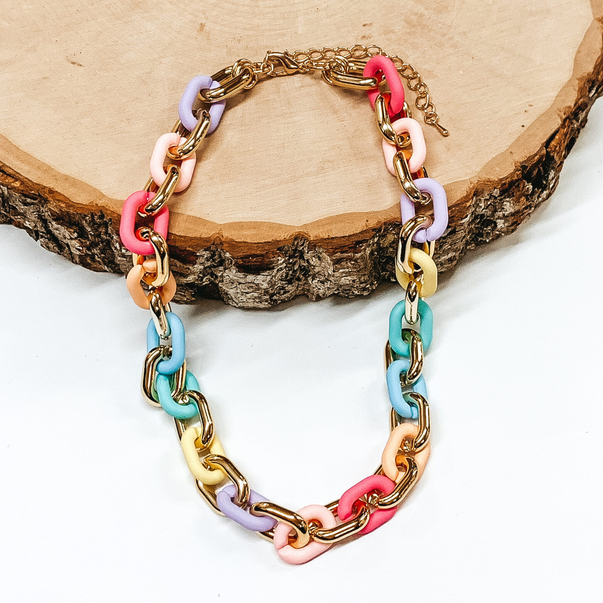 Thick chain link necklace. This necklace includes gold links and multicolored, matte links. This necklace is pictured partially laying on a piece of wood on a white background.