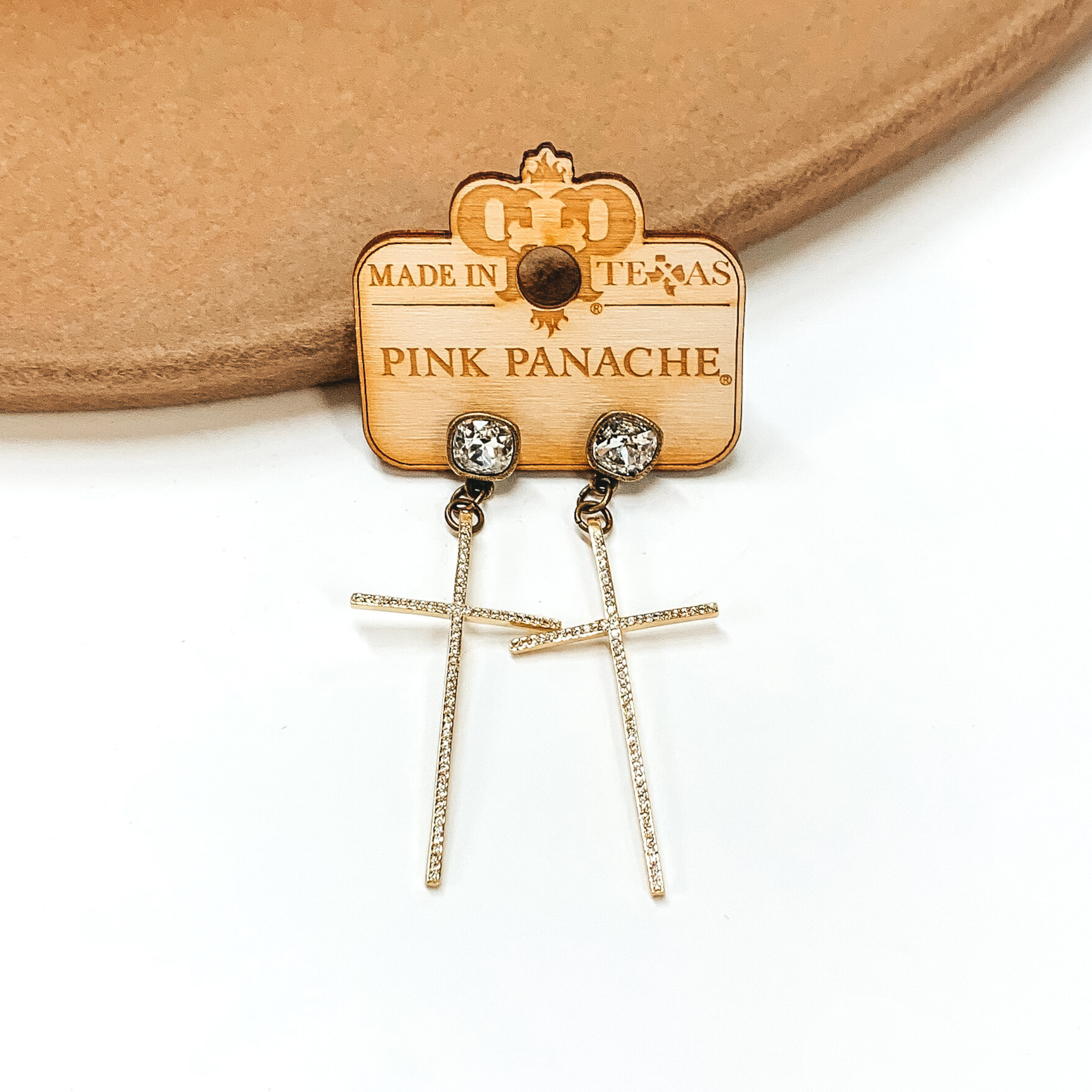 Clear cushion cut crystal studs in a bronze setting with a thin, gold hanging cross. These earrings are pictured on a white and tan background. 