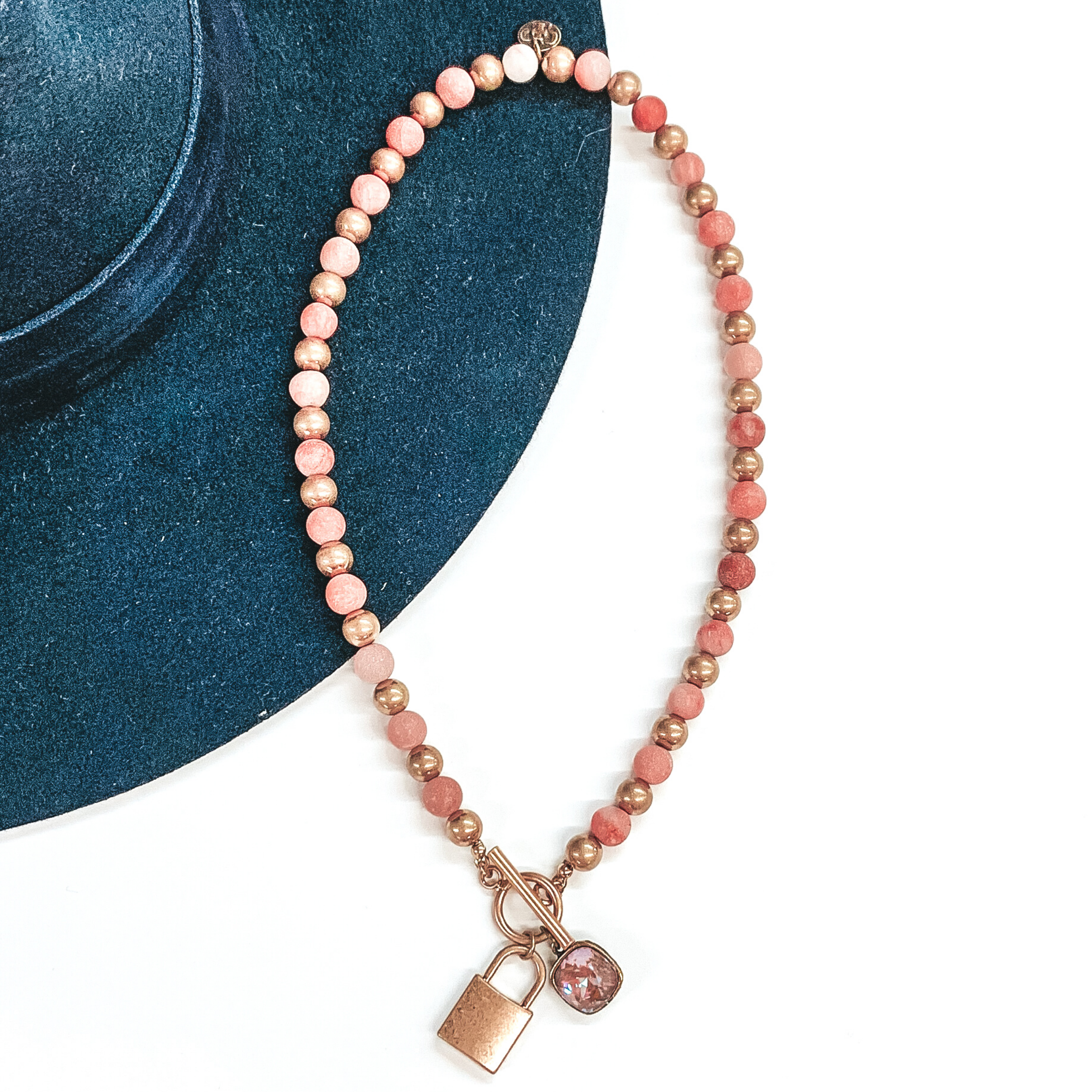 Bronze and pink colored beaded necklace with front toggle clasp. There is also a gold lock pendant and a hanging cappuccino delight cushion cut crystal. This necklace is pictured on a white and navy background.