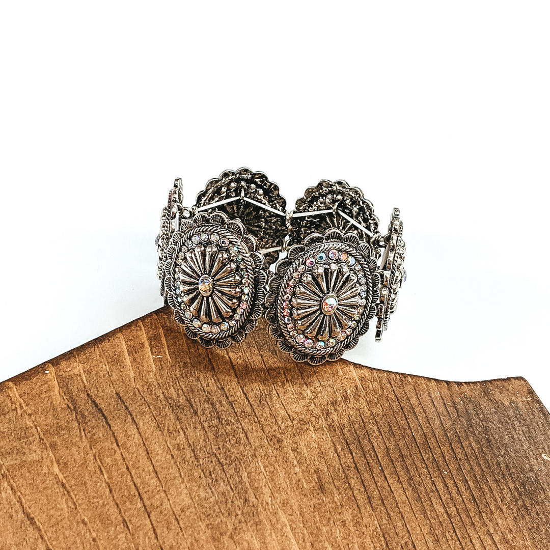 Silver oval conchos with ab crystals that form together in a stretchy bracelet. This bracelet is pictured laying partially on a piece of wood on a white background. 