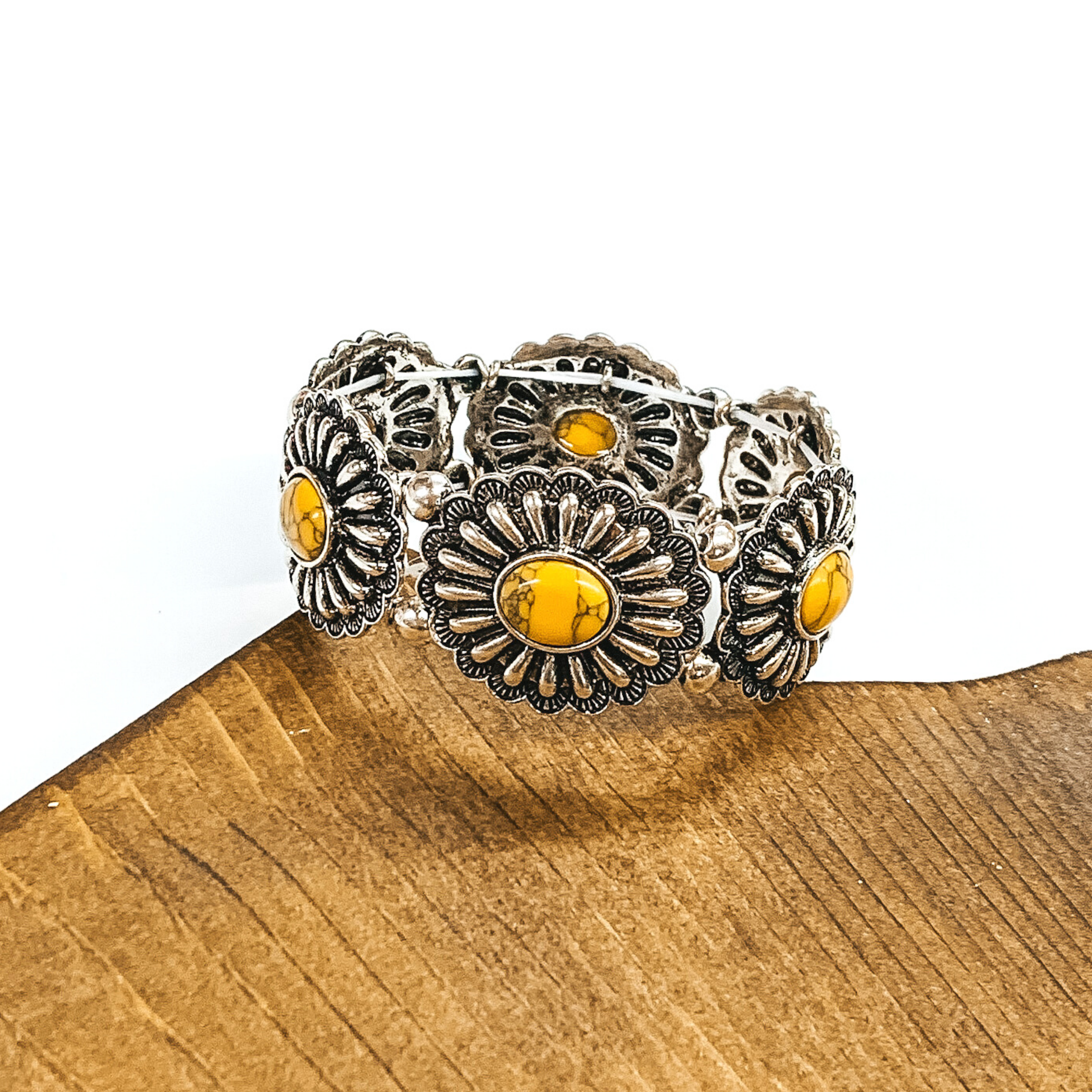 Silver, oval concho stretchy bracelet with oval center stones in yellow. This bracelet is pictured partially laying on a piece of wood and on a white background.