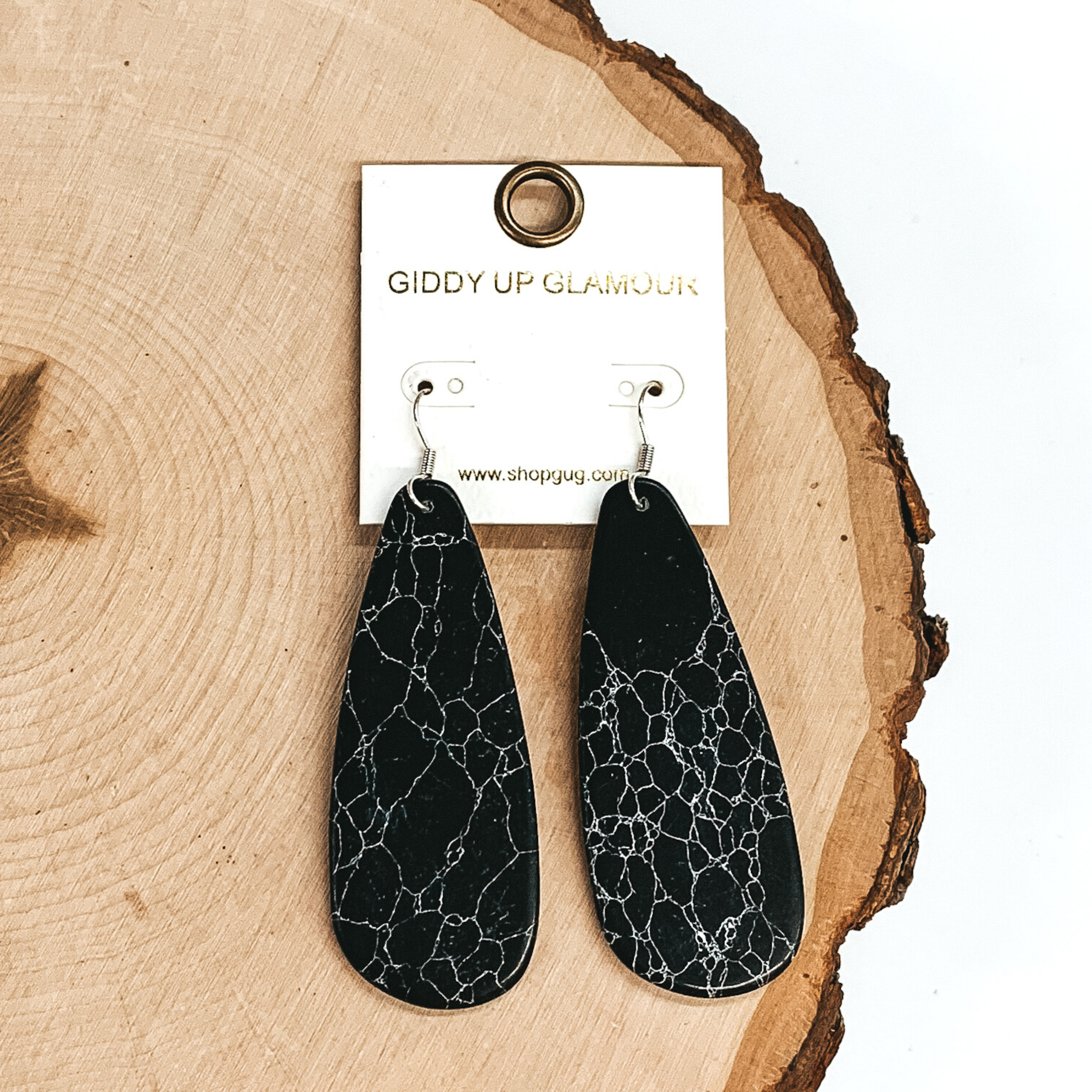 Teardrop shaped stone earrings. These dangle earrings have a marbled look and is in a black color. These earrings are pictured laying on a piece of wood on a white background.