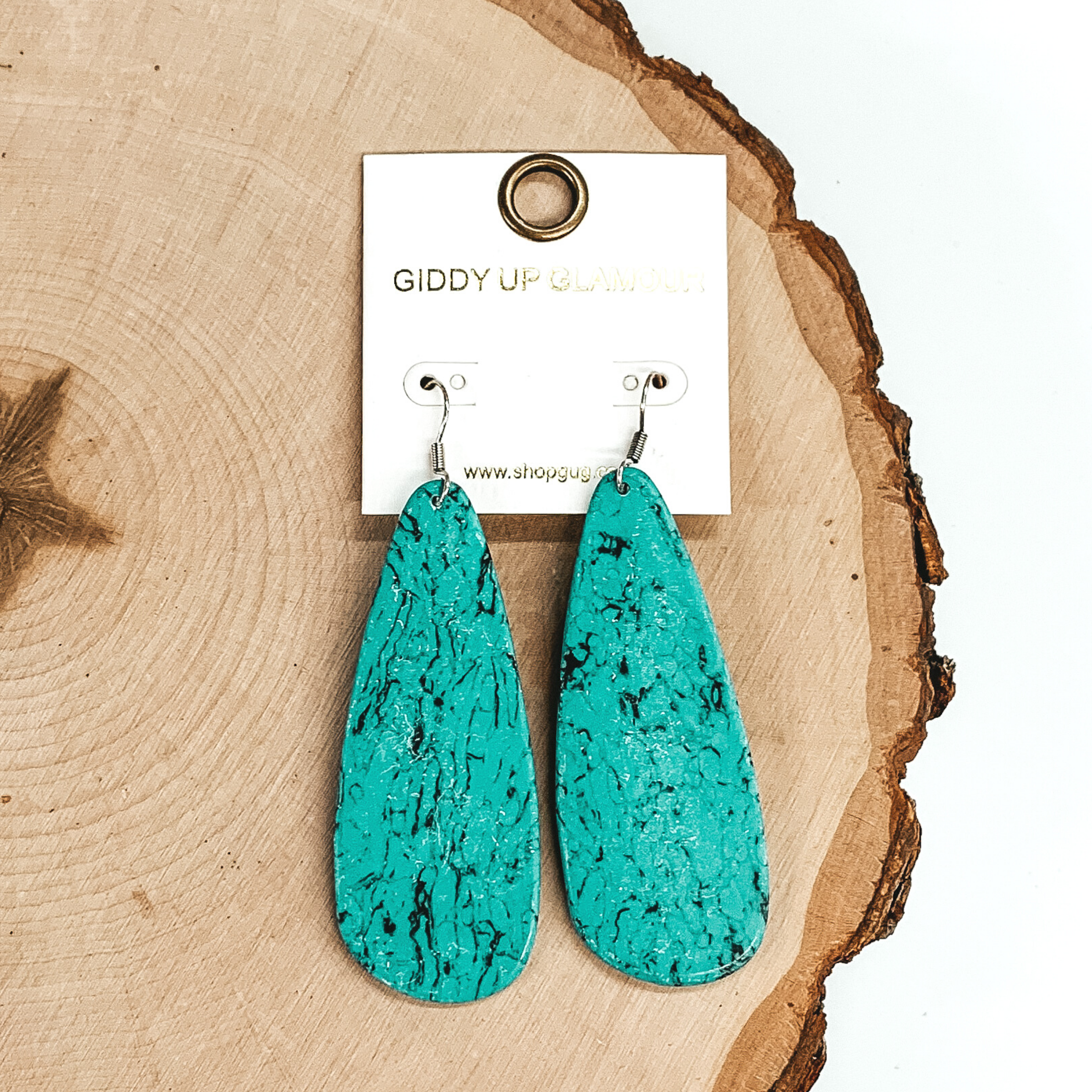 Teardrop shaped stone earrings. These dangle earrings have a marbled look and is in a turquoise color. These earrings are pictured laying on a piece of wood on a white background.