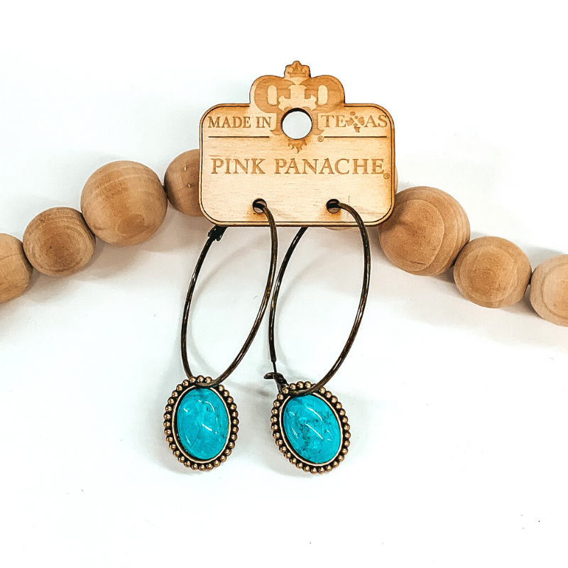 Large bronze hoop earrings with a hanging oval pendant. The pendant is outlined in bronze beads and has a center turquoise colored stone. These hoops are pictured in front of tan beads on a white background. 