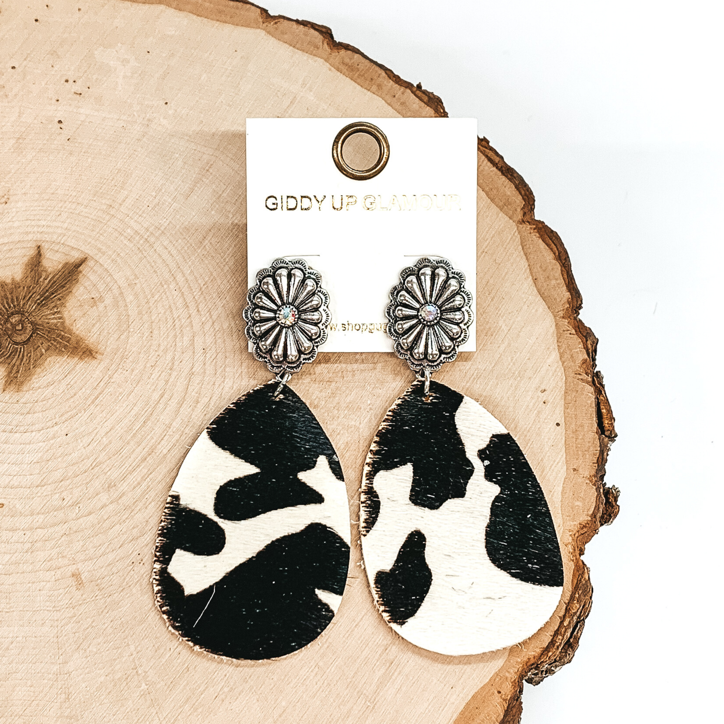 These earrings have small, oval, silver concho studs with a hanging teardrop pendant. This pendant has a white and black cow hide print. These earrings are pictured laying on a piece of wood on a white background. 
