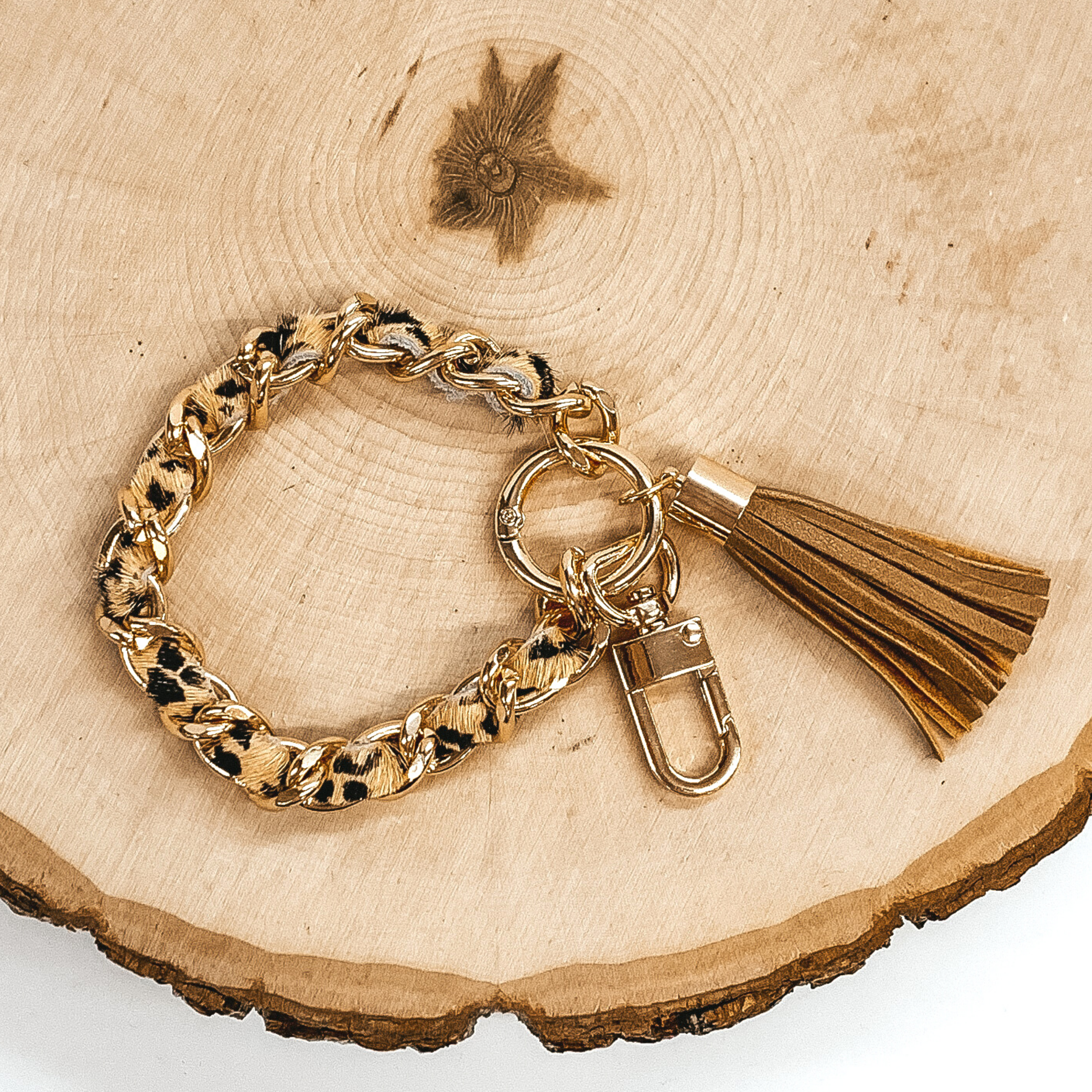Gold chain link bangle with a cowhide like material running through the link. The material is a black and tan color. This bangle has two key chains and a tan tassel. It is pictured on a piece of wood on a white background.