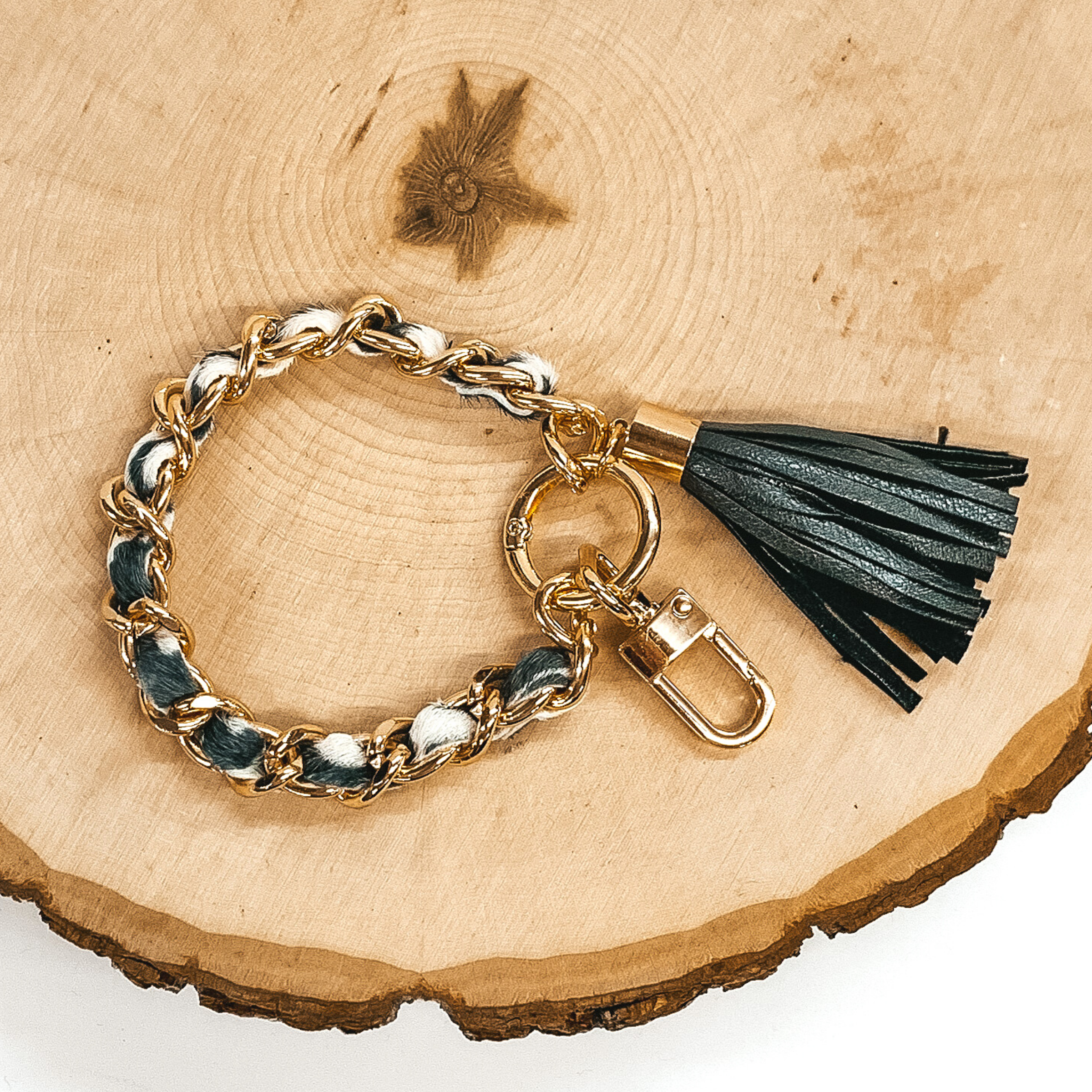 Gold chain link bangle with a cowhide like material running through the link. The material is a black and white color. This bangle has two key chains and a black tassel. It is pictured on a piece of wood on a white background.