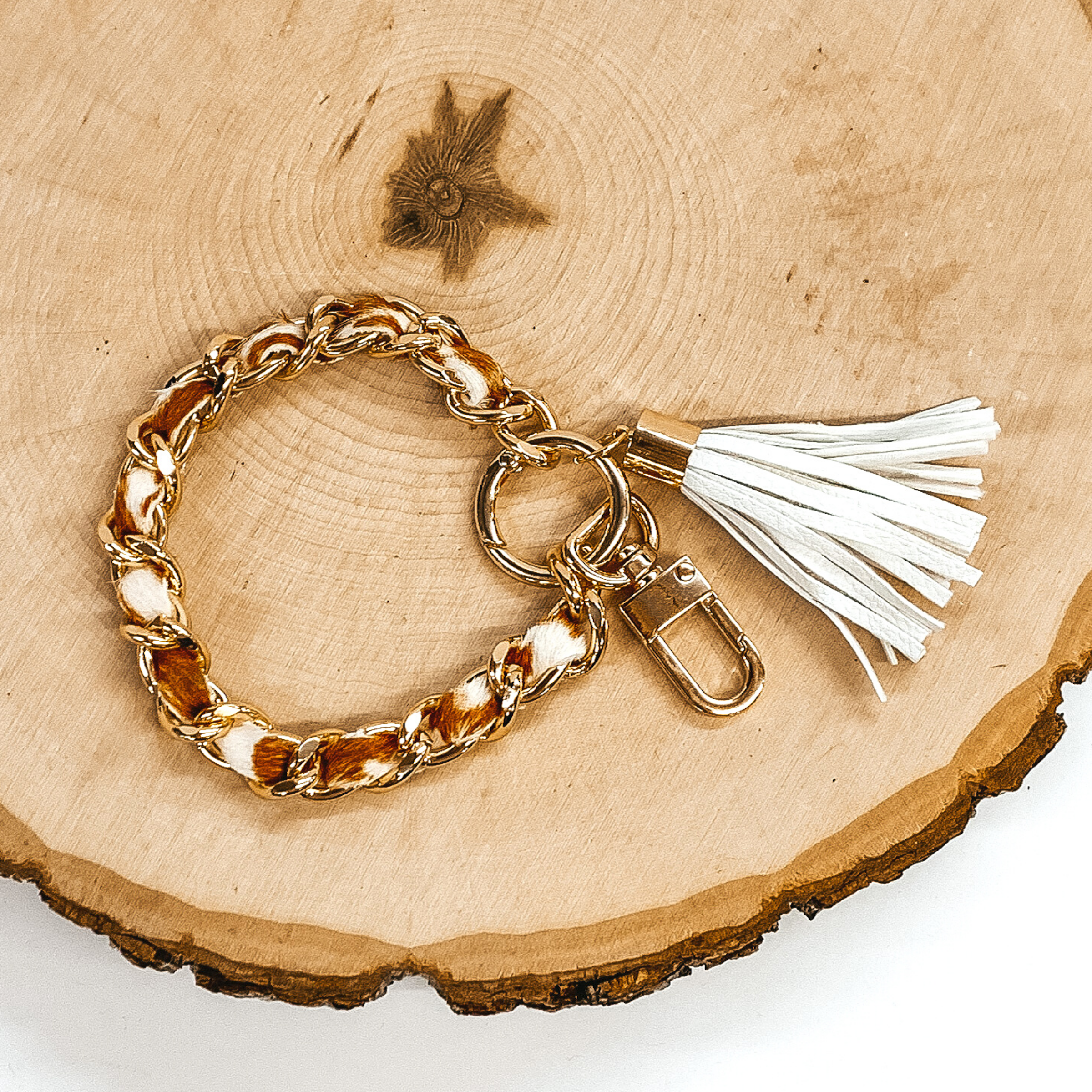 Gold chain link bangle with a cowhide like material running through the link. The material is a brown and white color. This bangle has two key chains and a white tassel. It is pictured on a piece of wood on a white background. 
