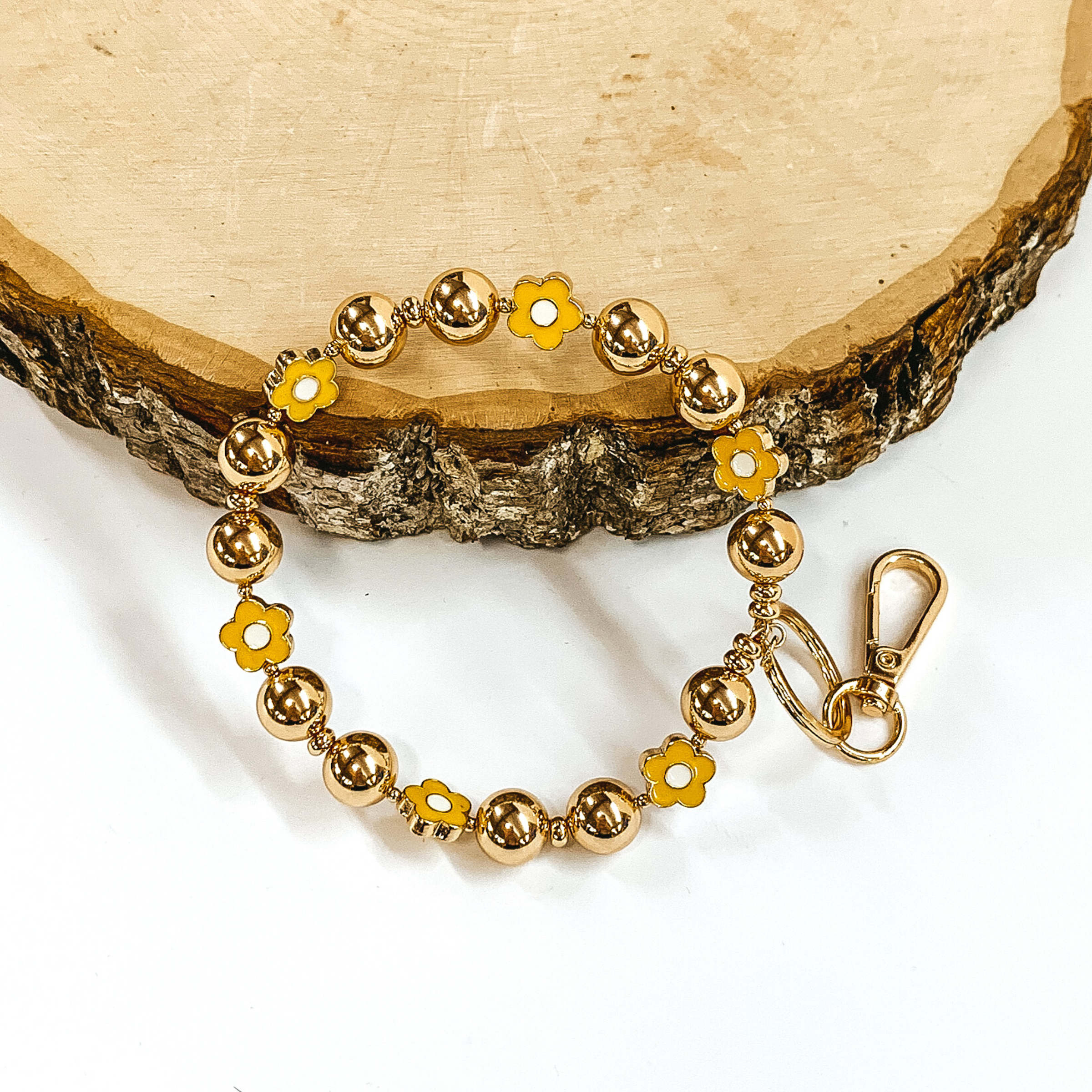 Gold beaded bracelet with yellow flower spacers. There is a gold key ring and clasp on one end of the bangle. It is pictured laying against a piece of wood on a white background.
