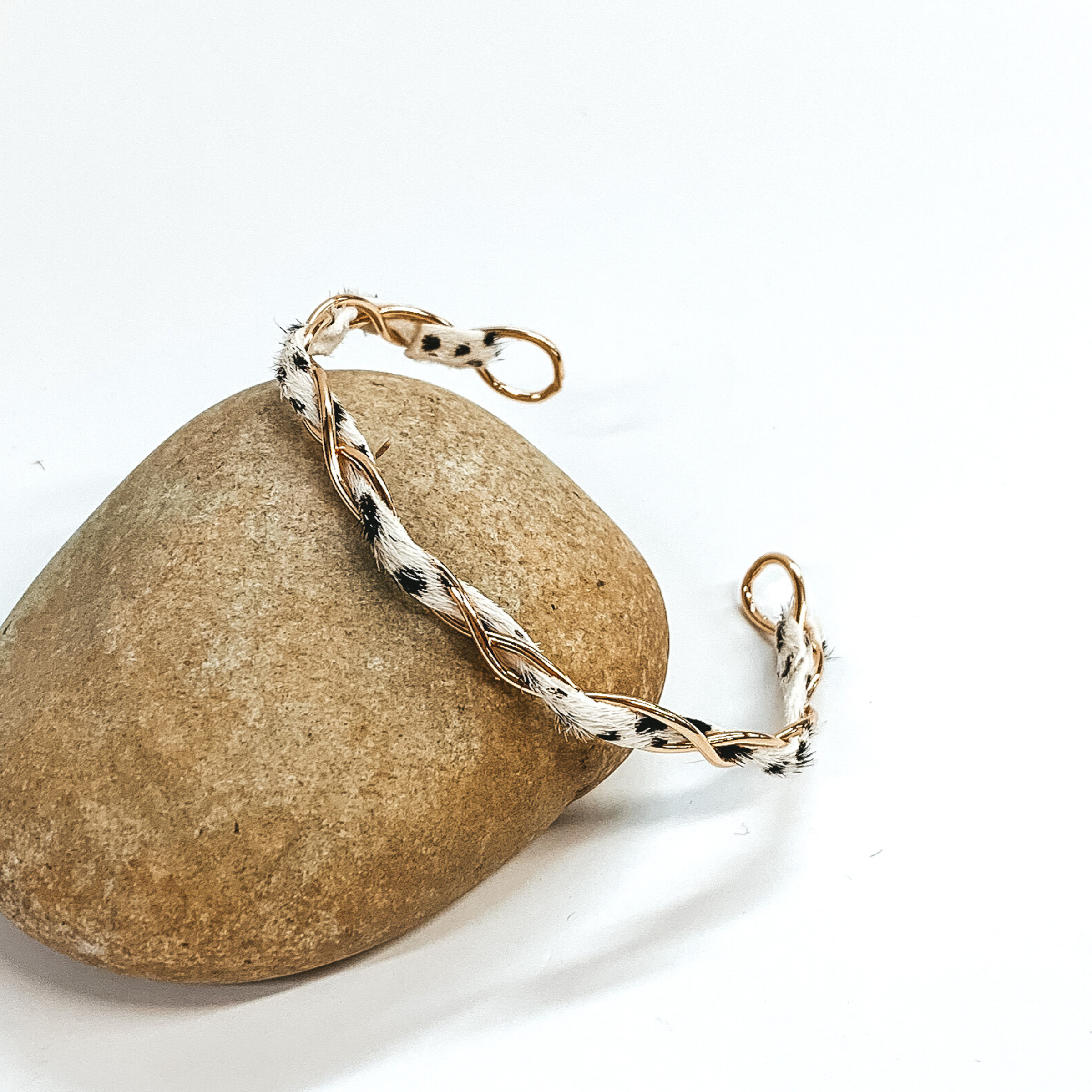 Gold twisted wire bracelet with faux fur inlay in a white dalmatian print. This bracelet is pictured laying against a rock on a white background.