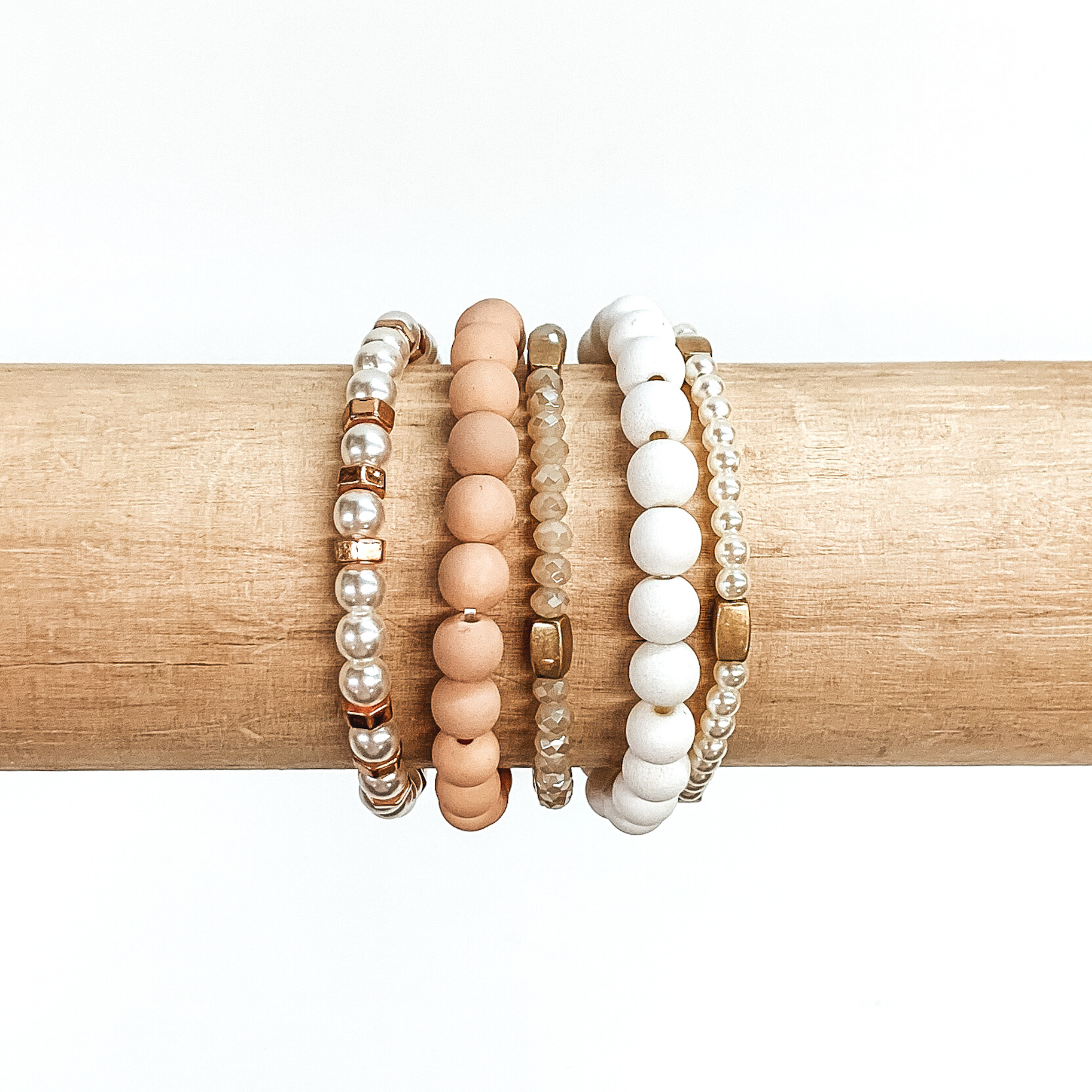 Beaded bracelet set with five different bracelets. Two are matte beaded bracelets with one in baby pink and one in white. Two bracelets include white pearls with gold or rose gold spacers throughout. The last bracelet has tan crystal beads with gold spacers. these bracelets are pictured on a wood bracelet holder on a white background. 