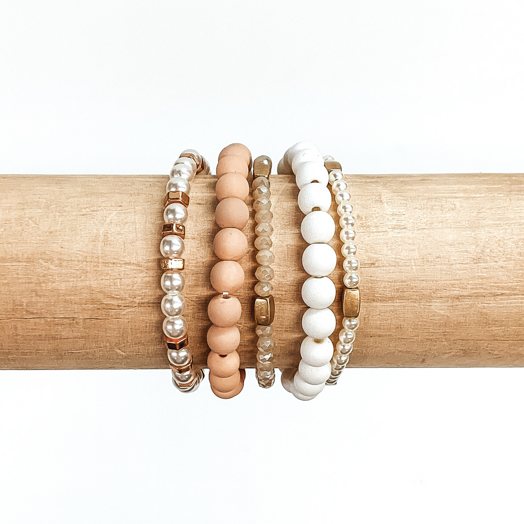Beaded bracelet set with five different bracelets. Two are matte beaded bracelets with one in baby pink and one in white. Two bracelets include white pearls with gold or rose gold spacers throughout. The last bracelet has tan crystal beads with gold spacers. these bracelets are pictured on a wood bracelet holder on a white background. 
