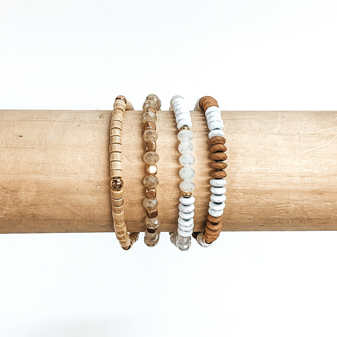 Beaded bracelet set with four different bracelets. Two of the bracelets include white, brown, or clear flat beads. One braclet has wood colored beads with gold spacers. The last bracelet has champagne crystal beads with gold spacers. These bracelets are pictured on a wood bracelet holder on a white background.