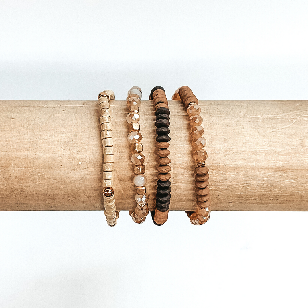 Beaded bracelet set with four different bracelets. Two of the bracelets include light brown, brown, or champagne flat beads. One braclet has wood colored beads with gold spacers. The last bracelet has champagne crystal beads with gold spacers. These bracelets are pictured on a wood bracelet holder on a white background.