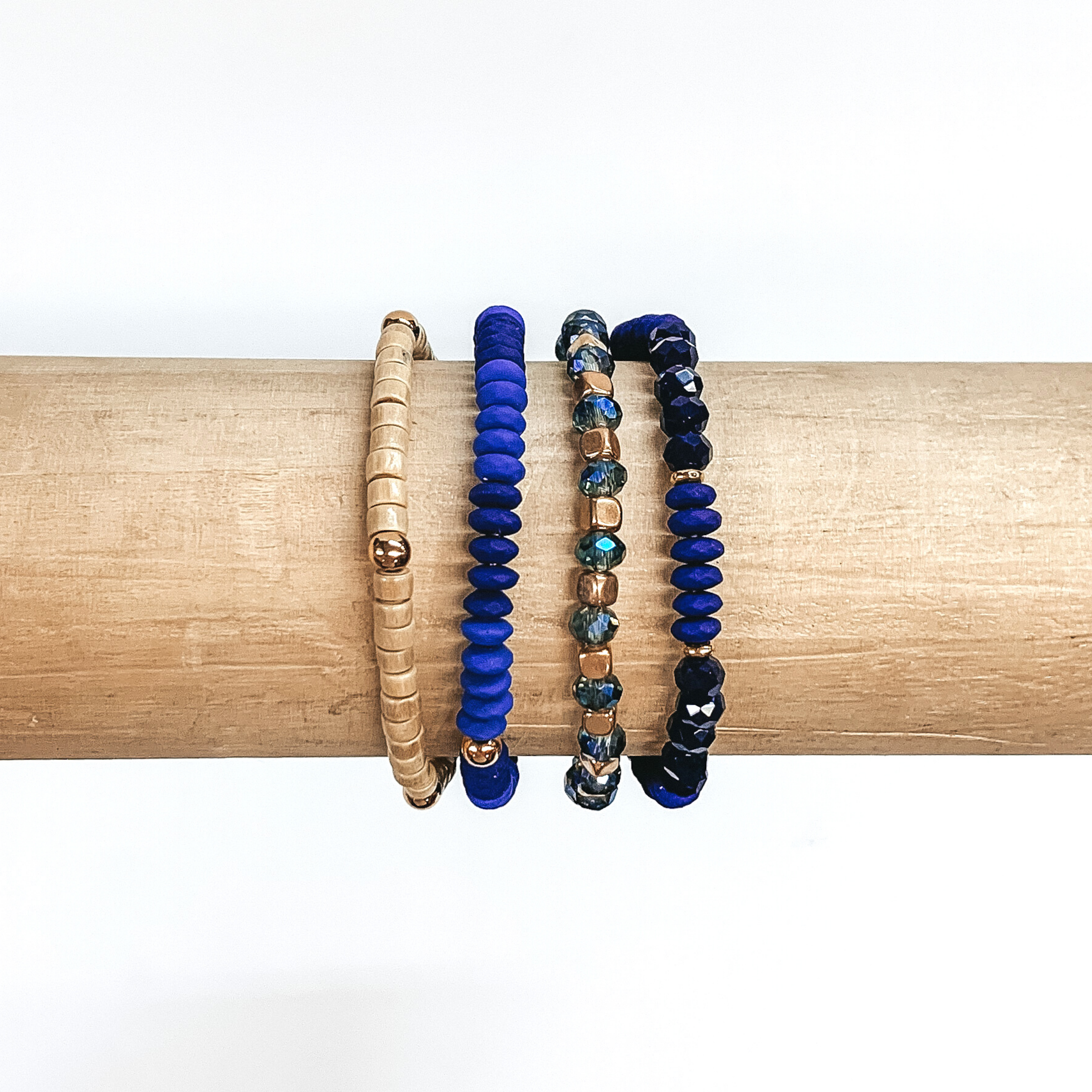 Beaded bracelet set with four different bracelets. Two of the bracelets include blue, navy, or cyrstal blue flat beads. One braclet has wood colored beads with gold spacers. The last bracelet has blue crystal beads with gold spacers. These bracelets are pictured on a wood bracelet holder on a white background.
