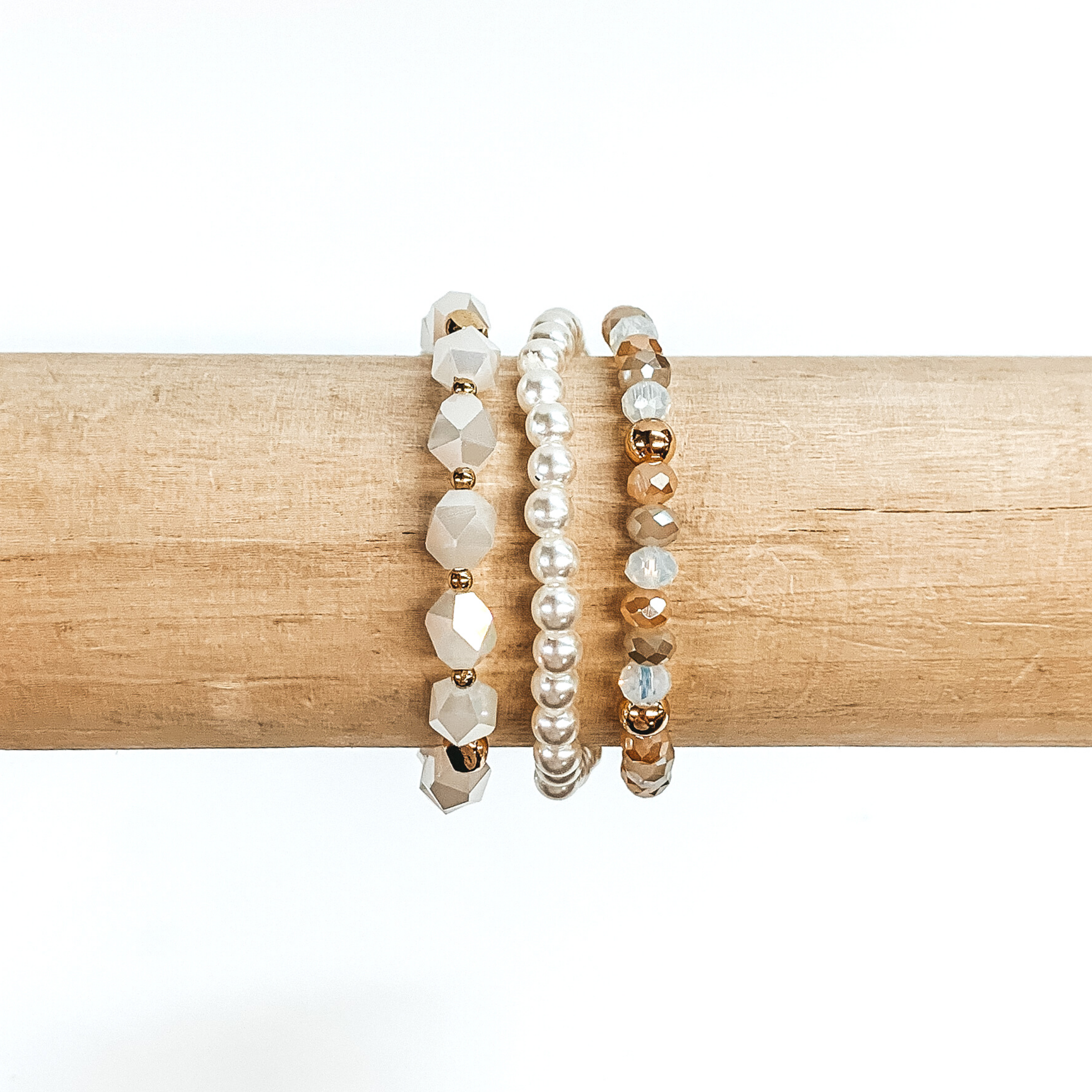 Beaded bracelet set with three different bracelets. One bracelet is white pearl beads only. The next one has big white crystal beads with gold spacers. The last braclet has a mixture of tan, champagne, and clear crystal beads with gold spacers. These bracelets are pictured on a wood bracelet holder on a white background.