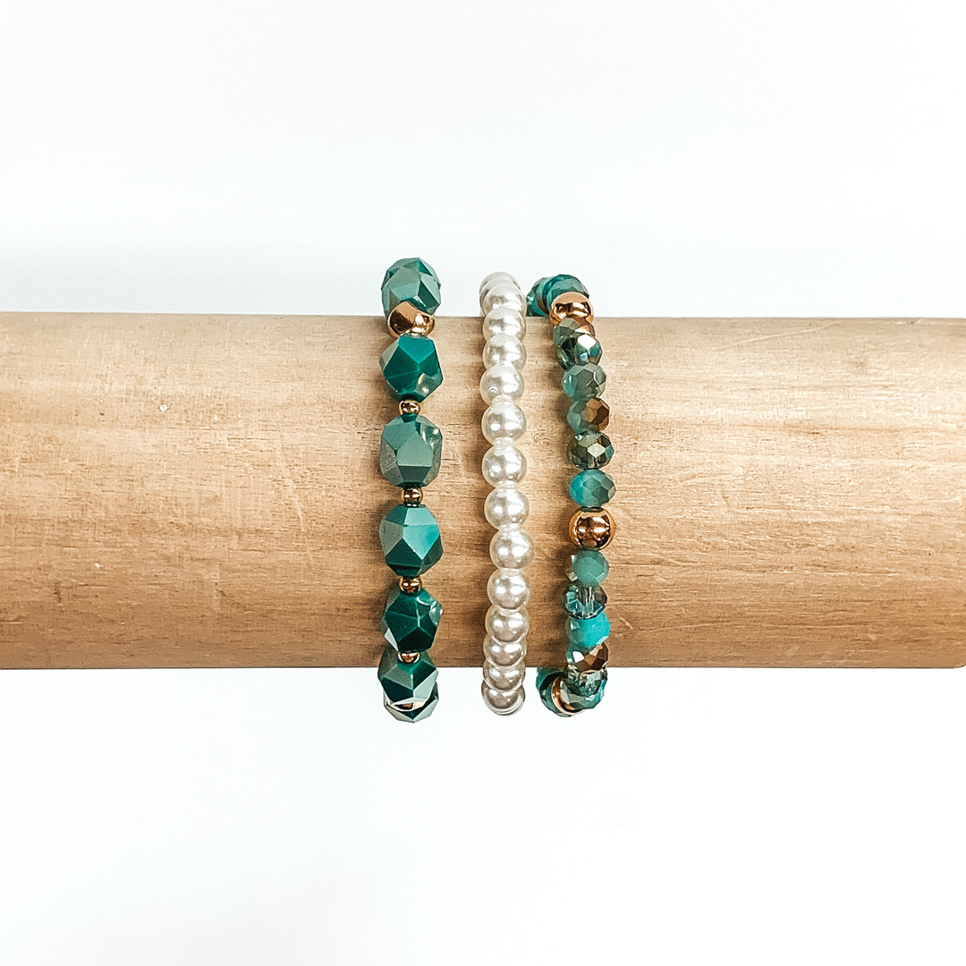 Beaded bracelet set with three different bracelets. One bracelet is white pearl beads only. The next one has big teal crystal beads with gold spacers. The last braclet has a mixture of tan and teal crystal beads with gold spacers. These bracelets are pictured on a wood bracelet holder on a white background.