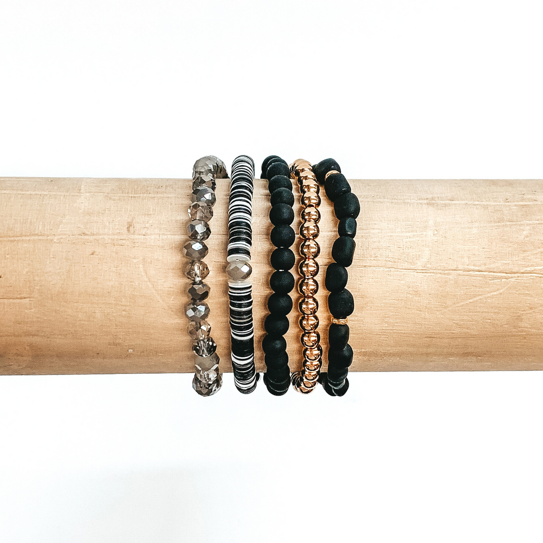 This is a beaded bracelet of five different bracelets. First, there is a plain gold beaded bracelet. One bracelet has black matte beads with regular round beads. Another has irregular shaped black matte beads with small gold spacers. There is a sequin beaded bracelet that has white and black colored sequins. The last bracelet has grey marbled crystal beaded bracelet. These bracelets are pictured on a wood bracelet holder on a white background.