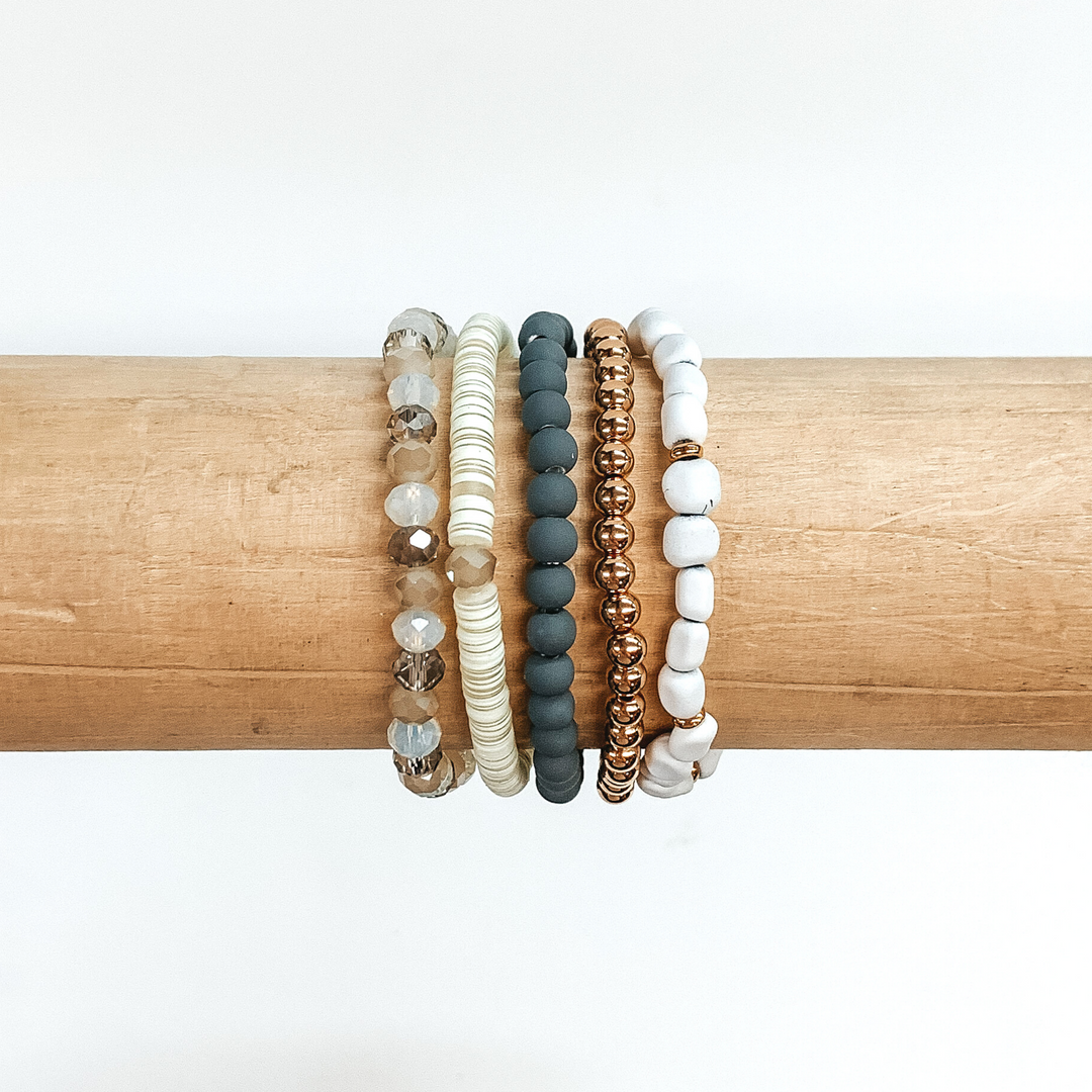 This is a beaded bracelet of five different bracelets. First, there is a plain gold beaded bracelet. One bracelet has grey matte beads with regular round beads. Another has irregular shaped white matte beads with small gold spacers. There is a sequin beaded bracelet that has white and beige colored sequins. The last bracelet has white, beige, and tan crystal beaded bracelet. These bracelets are pictured on a wood bracelet holder on a white background.