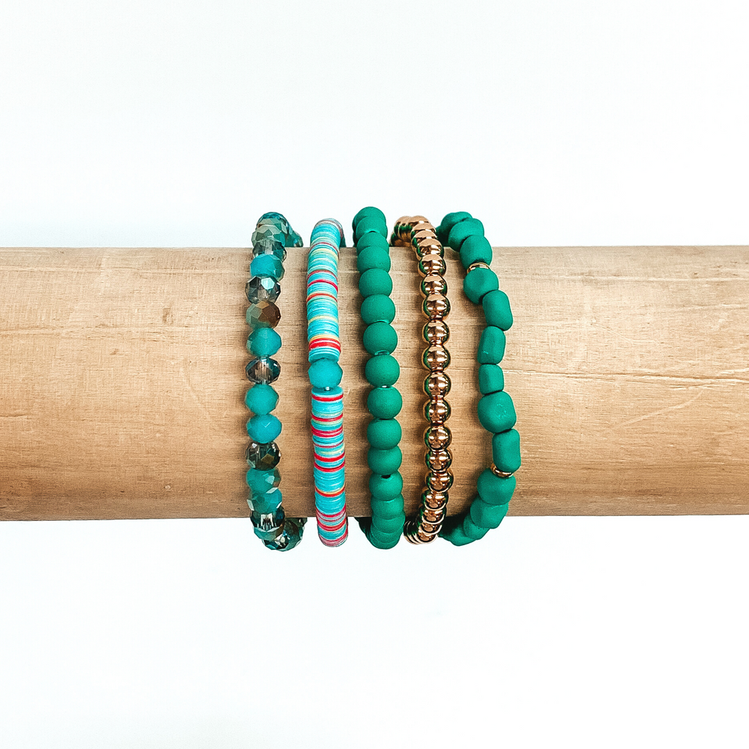This is a beaded bracelet of five different bracelets. First there is a plain gold beaded bracelet. Two bracelets has dark teal matte beads; one is regular round beads and the other has irregular shaped beads eith small gold spacers. There is a sequin beaded bracelet that has blue, red, and yellow colored sequins. The last bracelet is a teal marbled crystal beaded bracelet. These bracelets are pictured on a wood bracelet holder on a white background.