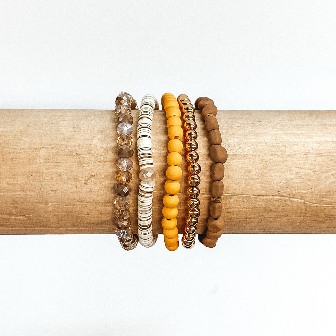 This is a beaded bracelet of five different bracelets. First, there is a plain gold beaded bracelet. One bracelet has yellow matte beads with regular round beads. Another has irregular shaped tan matte beads with small gold spacers. There is a sequin beaded bracelet that has white and tan colored sequins. The last bracelet has tan and beige marbled crystal beaded bracelet. These bracelets are pictured on a wood bracelet holder on a white background.