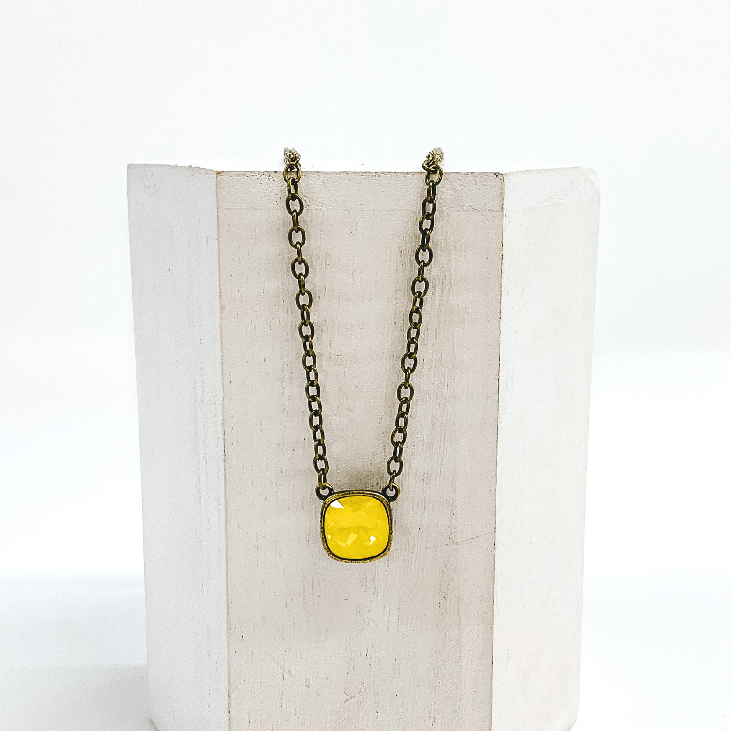 Bronze chained necklace with a square pendant with a yellow cushion cut crystal. This necklace is pictured laying on a white block on a white background. 