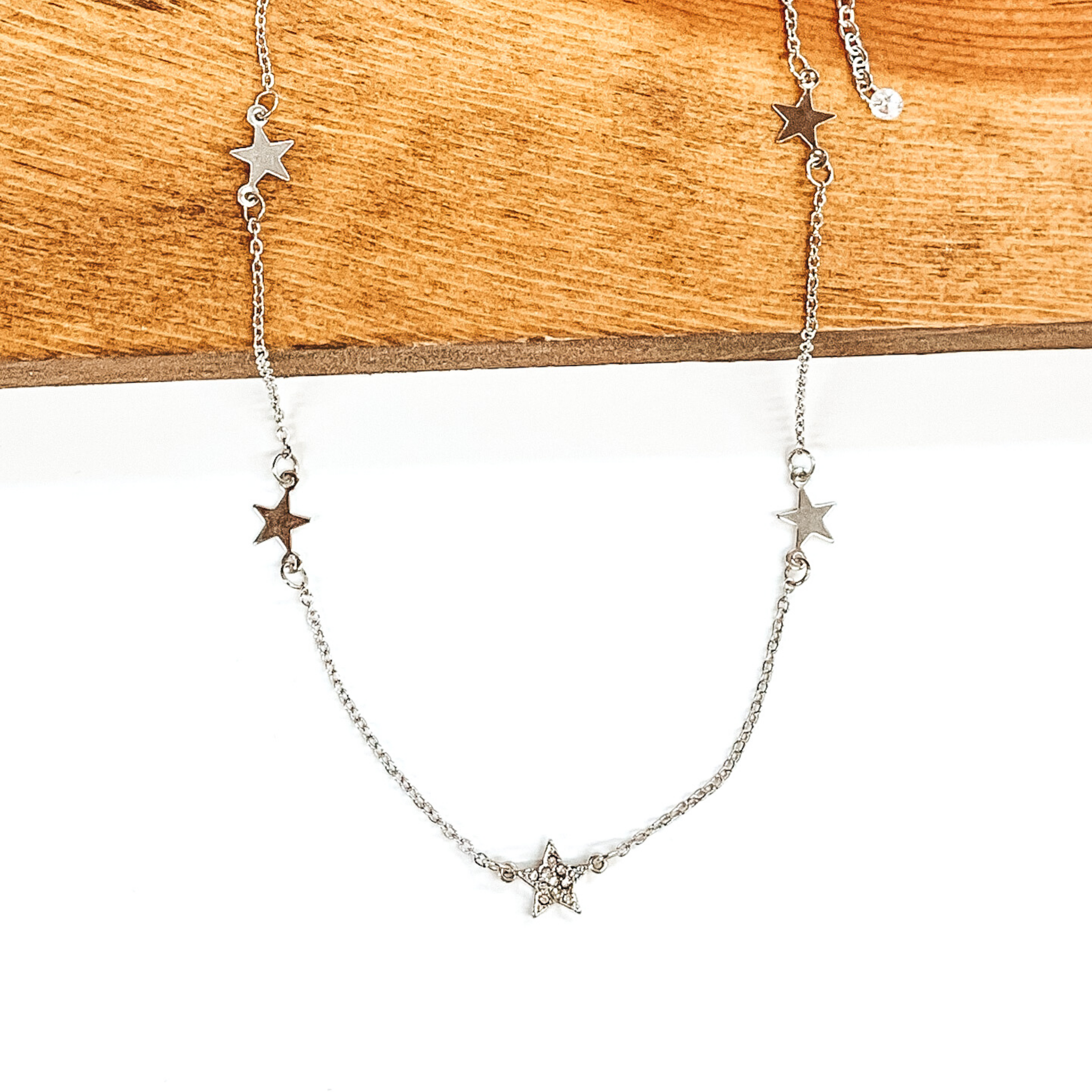 Silver chained necklace with silver star spacers. The middle star spacer has clear crystal on it. This necklace is pictured laying partially on a piece of wood on a white background. 