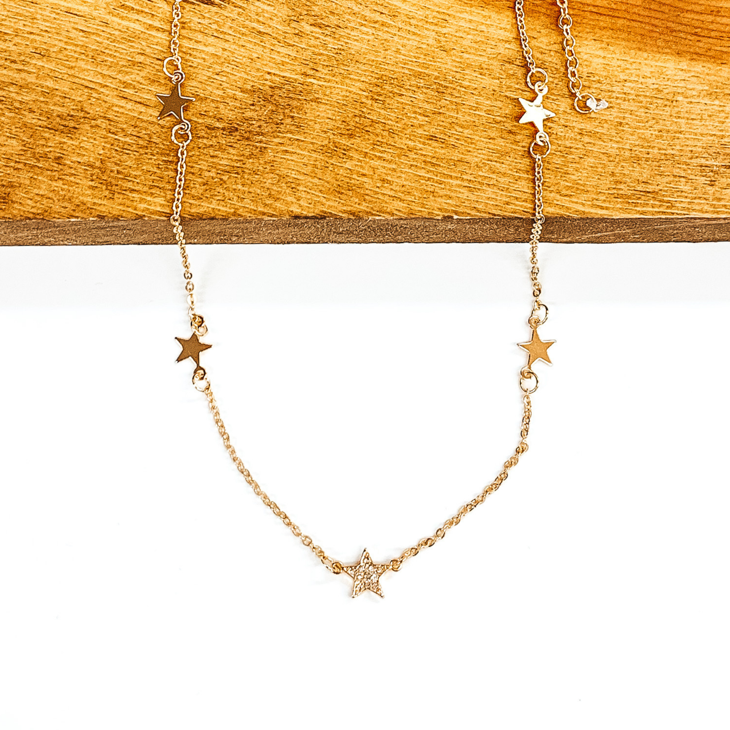 Gold chained necklace with gold star spacers. The middle star spacer has clear crystal on it. This necklace is pictured laying partially on a piece of wood on a white background.