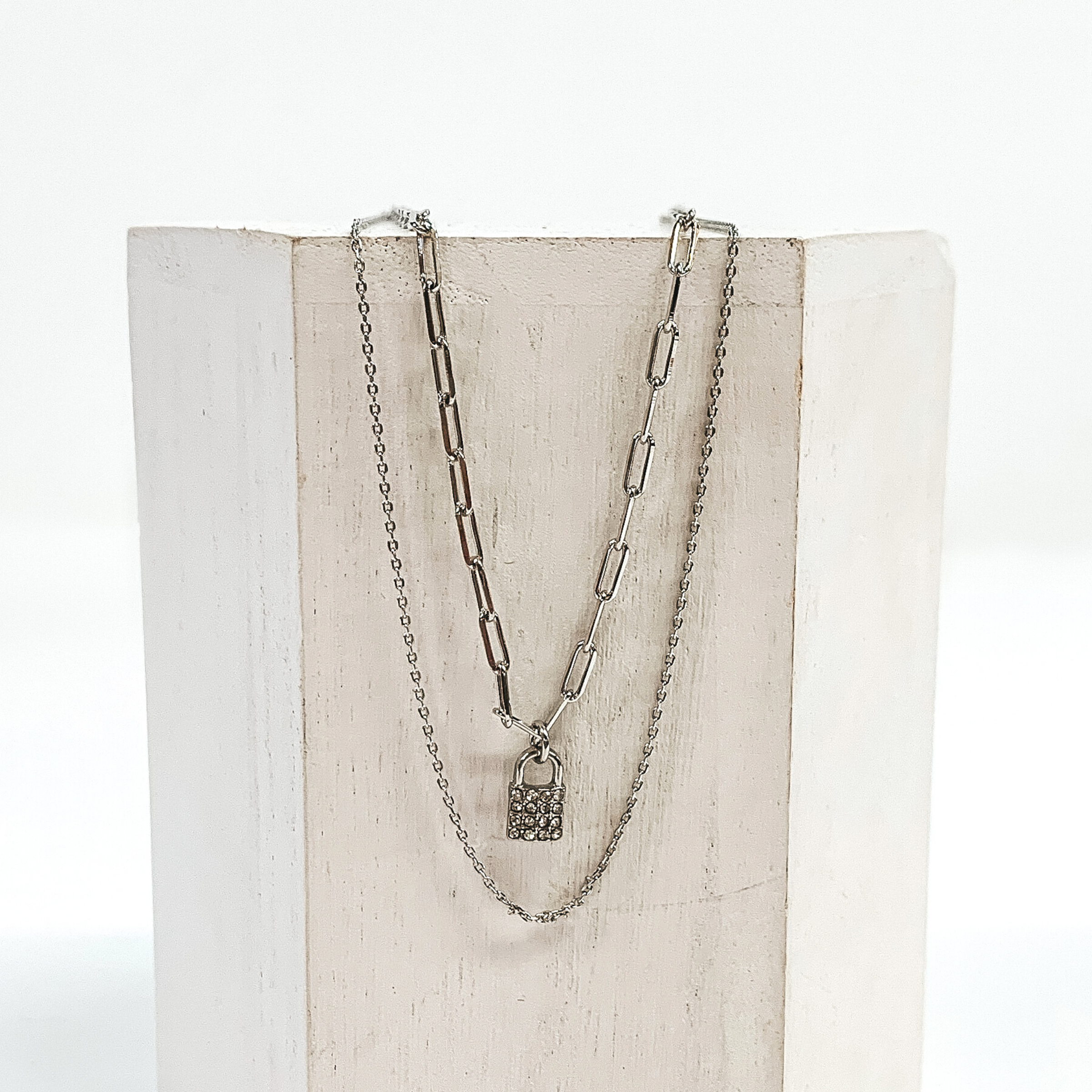 Silver, double stranded necklace. The longer chain is a plain silver chain. The shorter paperclip chain has a crystalized lock charm. This necklace is pictured laying on a white block on a white background.