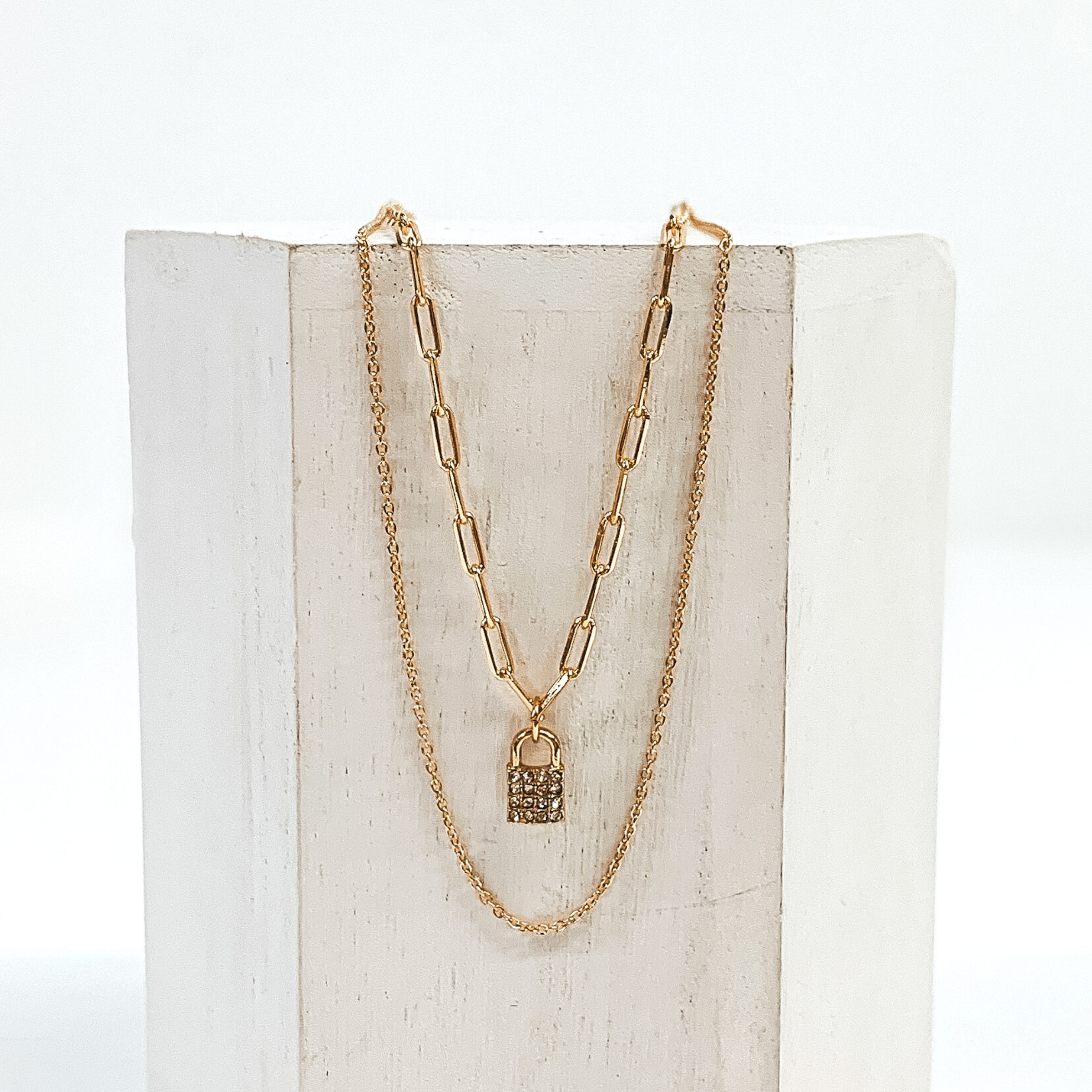 Gold, double stranded necklace. The longer chain is a plain gold chain. The shorter paperclip chain has a crystalized lock charm. This necklace is pictured laying on a white block on a white background. 