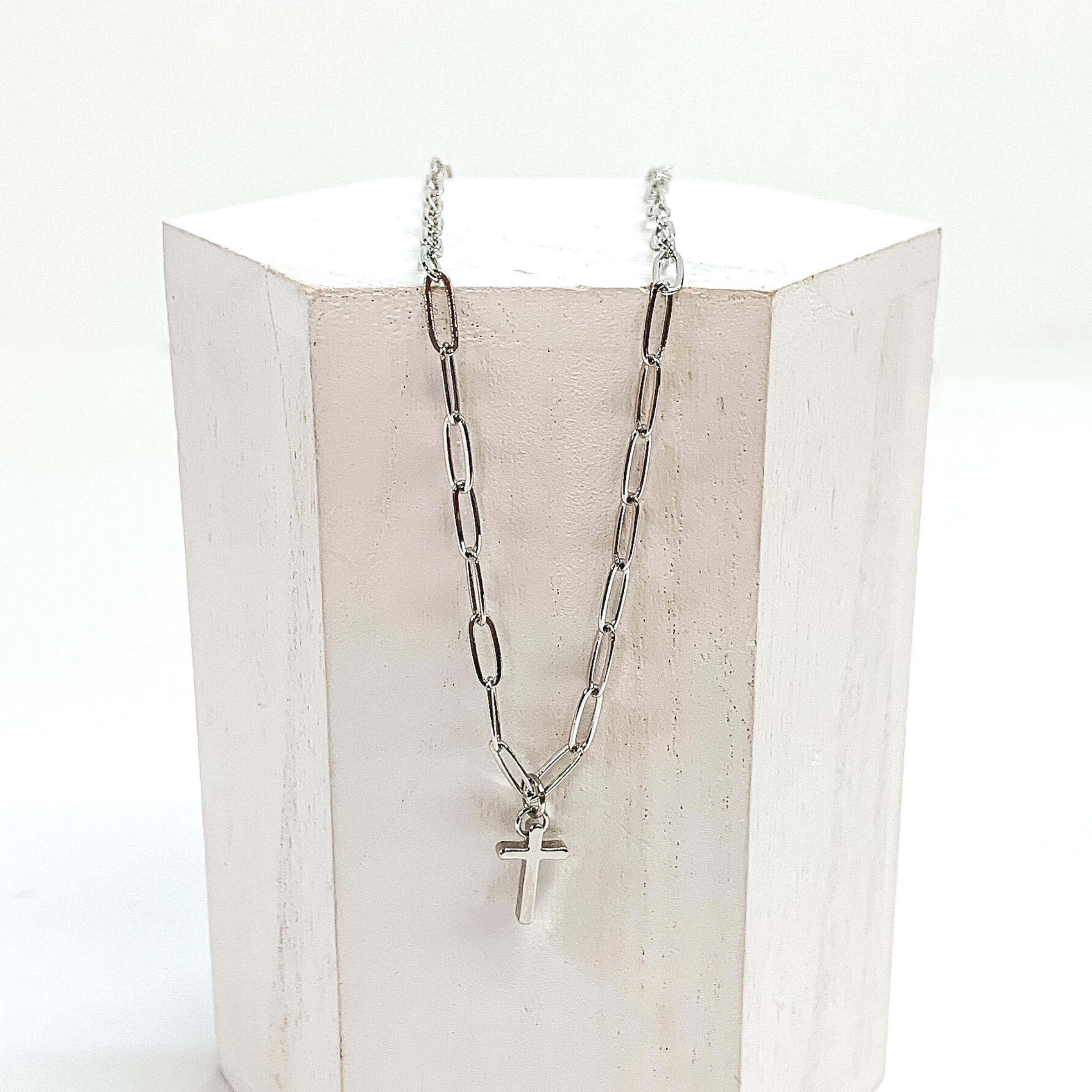 Silver paper clip chained necklace with a small, silver cross charm. This necklace is pictured laying on a white block on a white background.