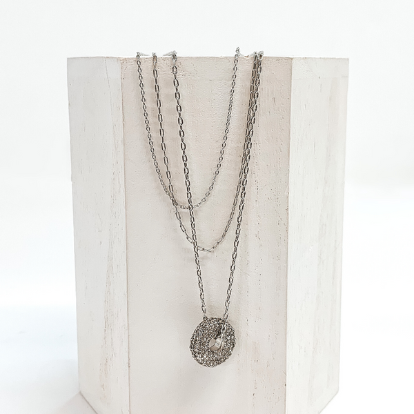 Silver, three stranded chained necklace with a round silver pendant. The pendant is covered in crystals. This necklace is pictured laying on a white block on a white background.