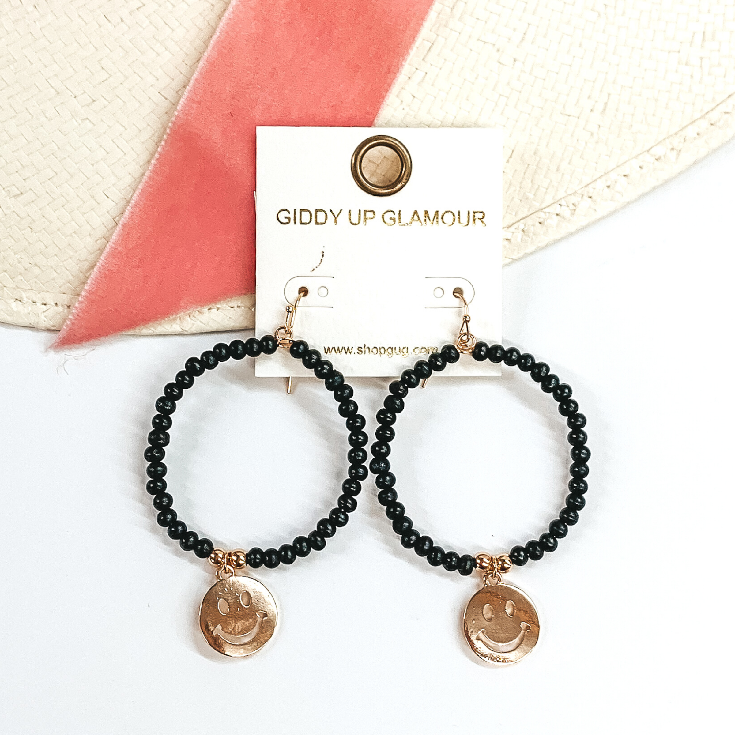 Circle drop earrings covered in black beads. These is a hanging smiley face charm in gold. These earrings are picture laying partially on a straw hat and pink velvet strip on a white background. 