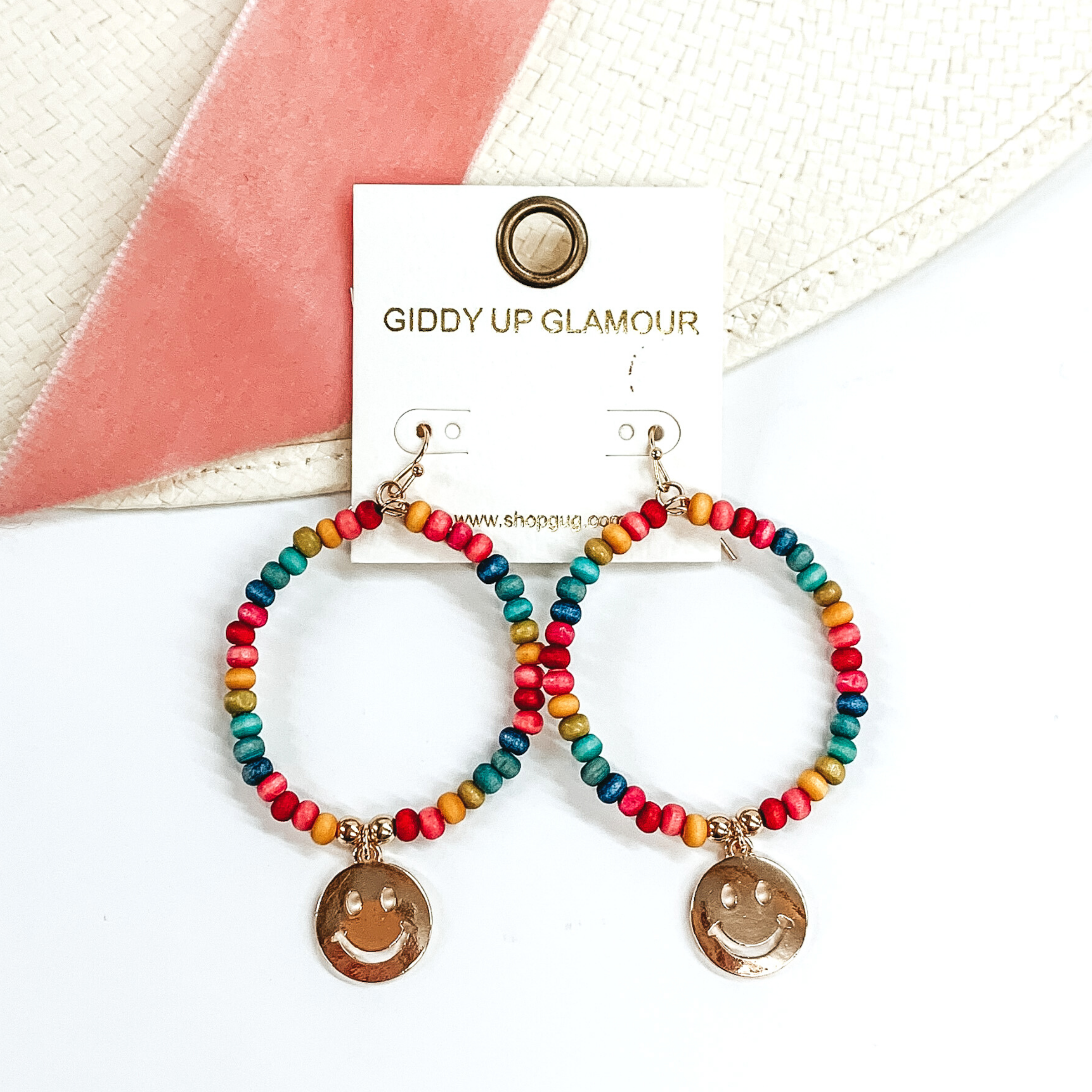 Circle drop earrings covered in multicolored beads. These is a hanging smiley face charm in gold. These earrings are picture laying partially on a straw hat and pink velvet strip on a white background.