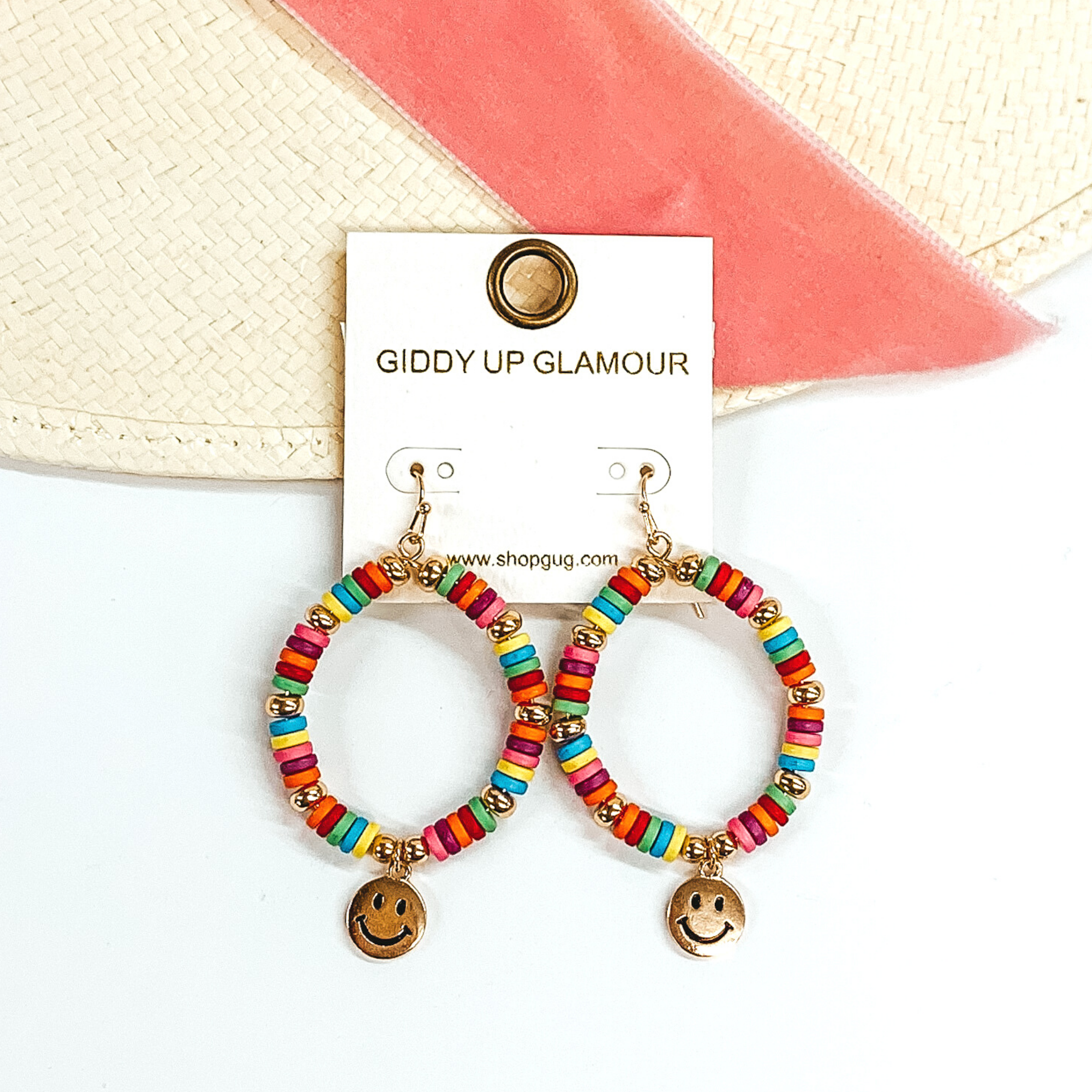 Hanging teardrop earrings with hanging gold smiley faces. These earrings are covered in multicolored and gold beads. These earrings are pictured laying partially on a straw hat with a pink velvet strip on a white background.