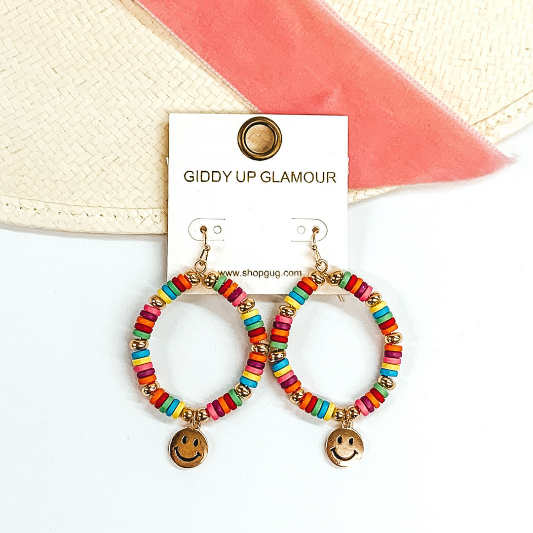 Hanging teardrop earrings with hanging gold smiley faces. These earrings are covered in multicolored and gold beads. These earrings are pictured laying partially on a straw hat with a pink velvet strip on a white background.