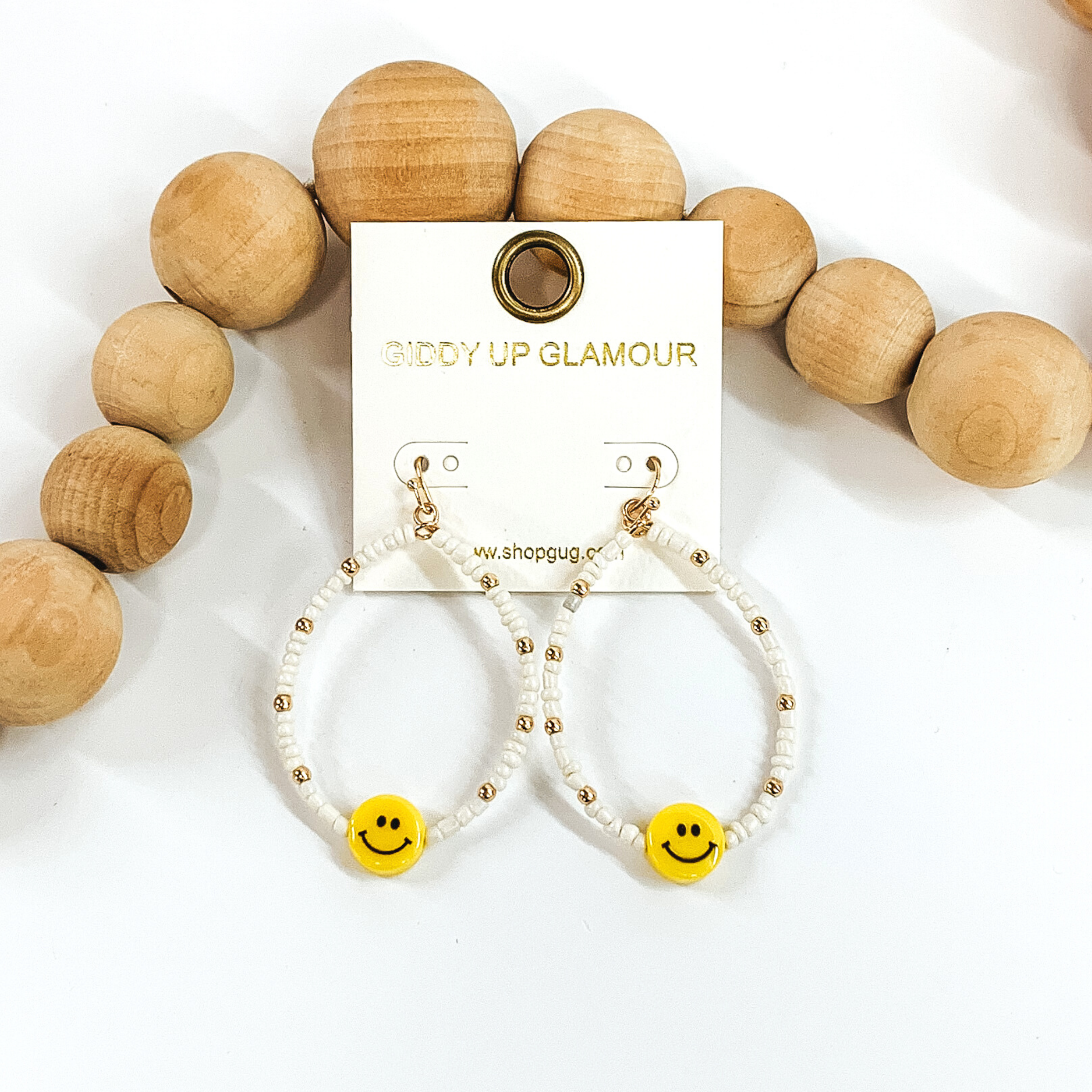 Hanging teardrop earrings with hanging yellow smiley faces. These earrings are covered in white beads with gold spacers. These earrings are pictured laying next to some tan beads on a white background.