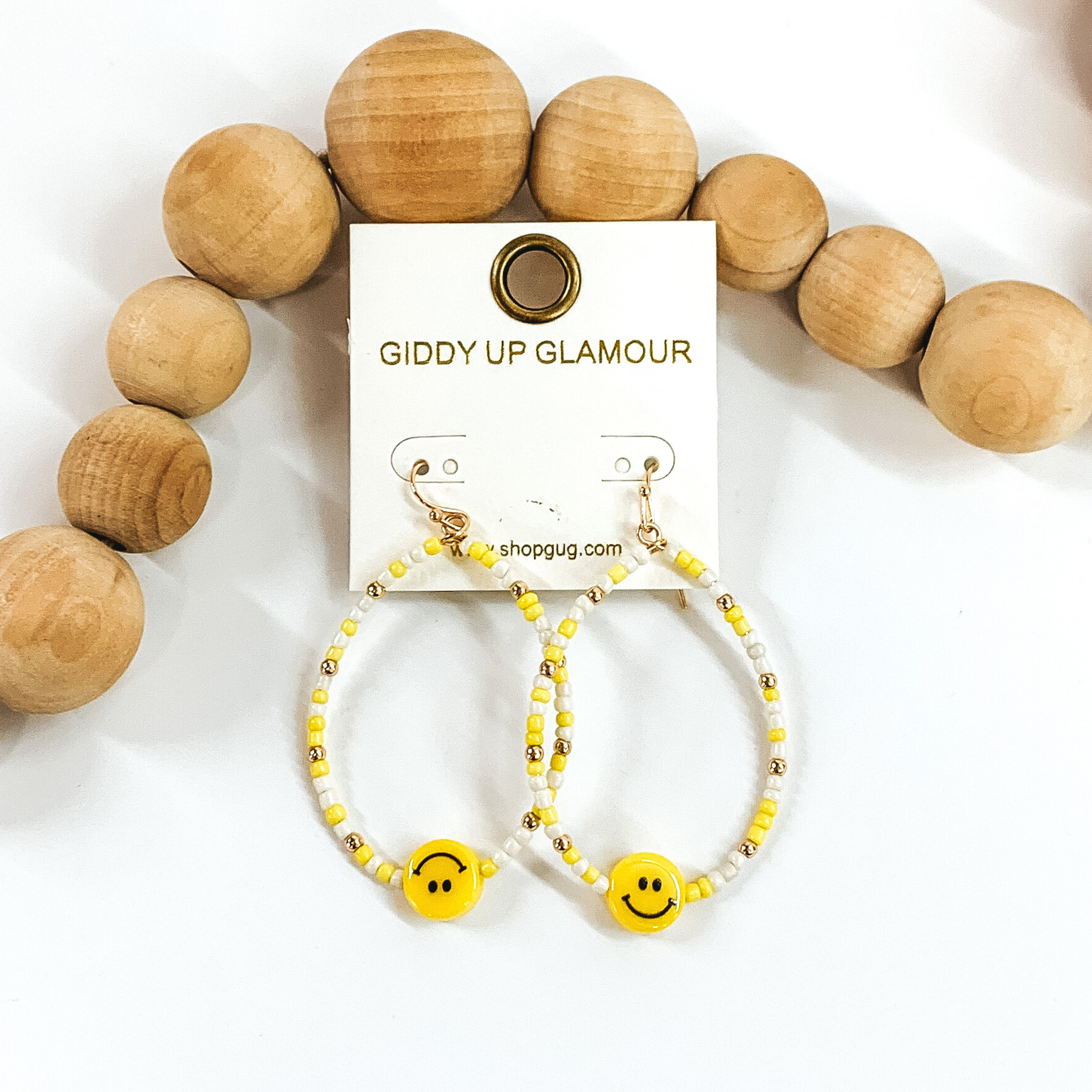 Hanging teardrop earrings with hanging yellow smiley faces. These earrings are covered in white and yellow beads with gold spacers. These earrings are pictured laying next to some tan beads on a white background.