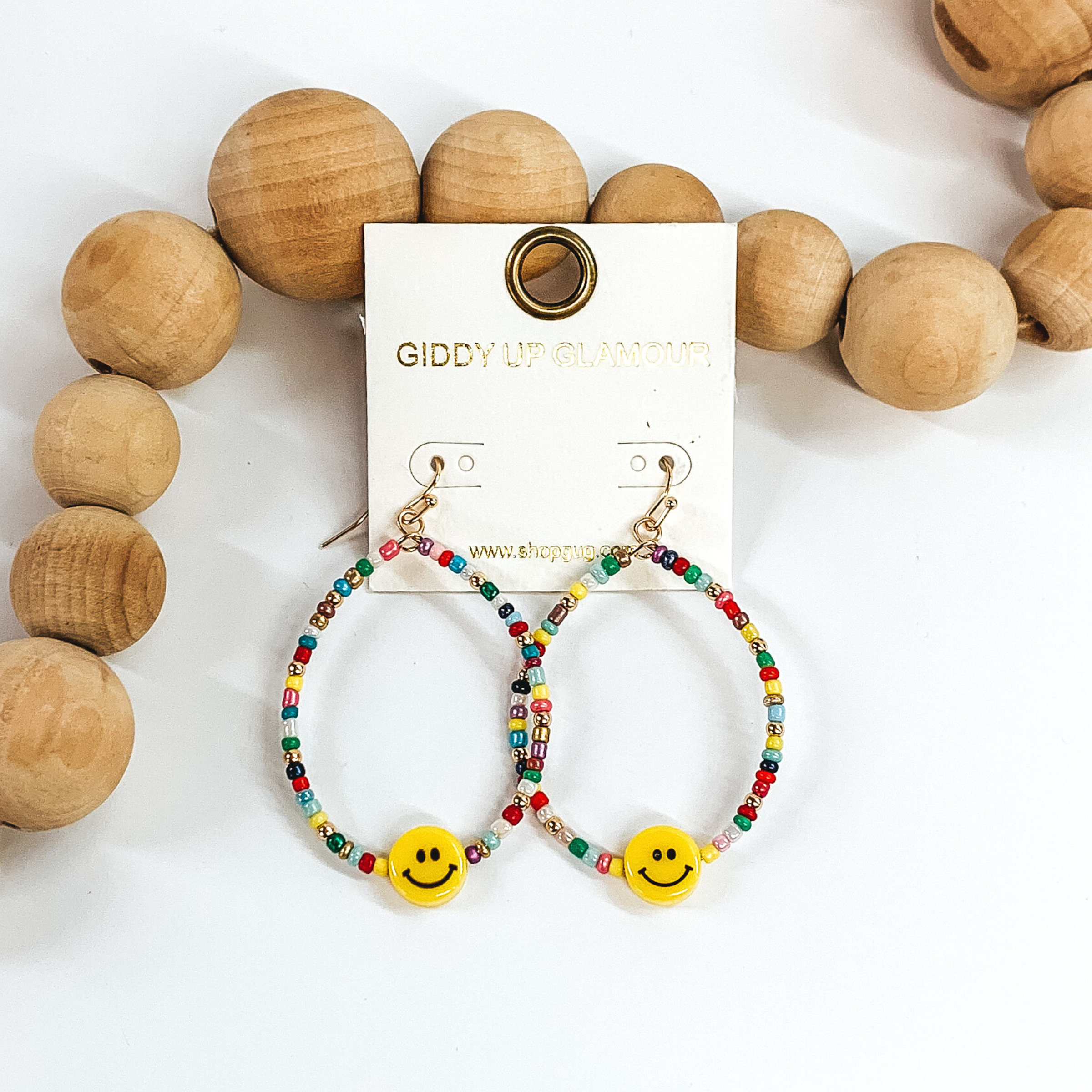Hanging teardrop earrings with hanging yellow smiley faces. These earrings are covered in multicolored beads with gold spacers. These earrings are pictured laying next to some tan beads on a white background.