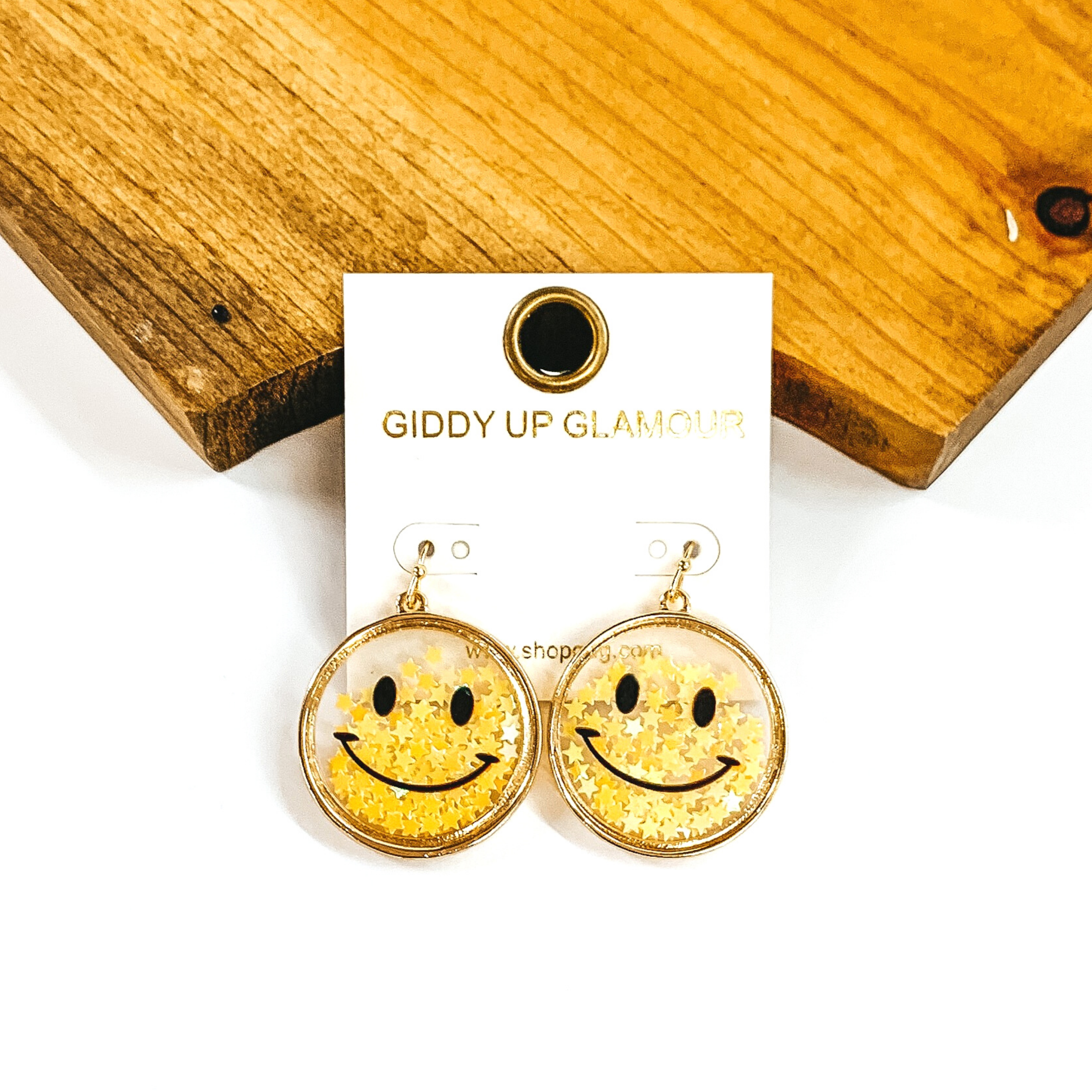 Hanging circle drop earrings with a smiley face. The circles are a small, clear shadow box with a gold outline. It is filled with tiny, yellow iridescent stars. these earrings are pictured in front of a dark piece of wood on a white background. 