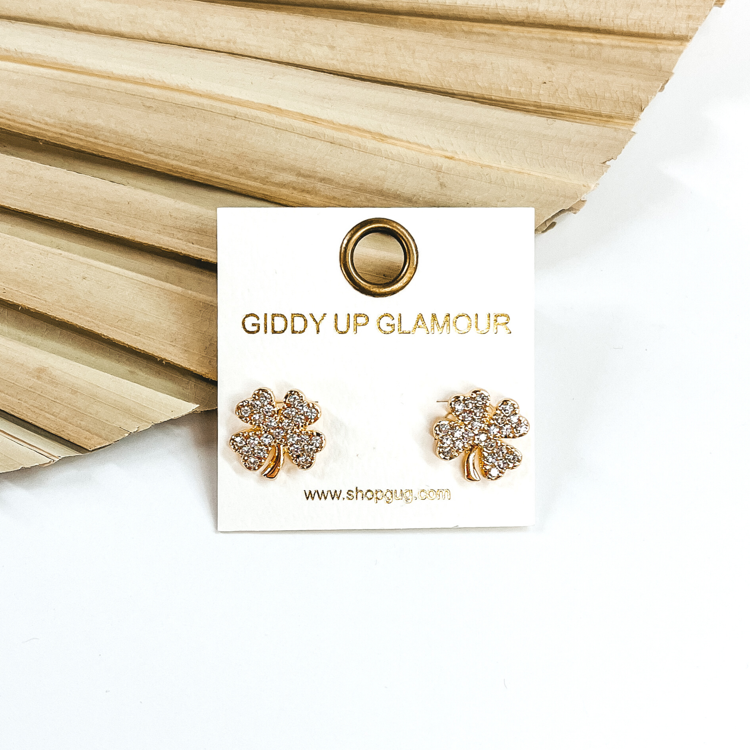 Gold four leaf clover stud earrings that are covered in clear crystals. These studs are pictured in front of a dried leaf on a white background.