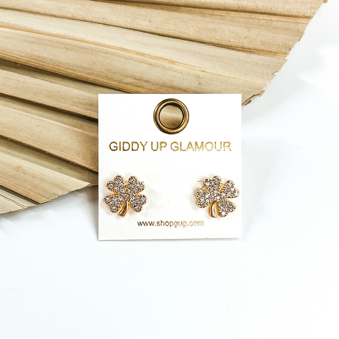 Gold four leaf clover stud earrings that are covered in clear crystals. These studs are pictured in front of a dried leaf on a white background.
