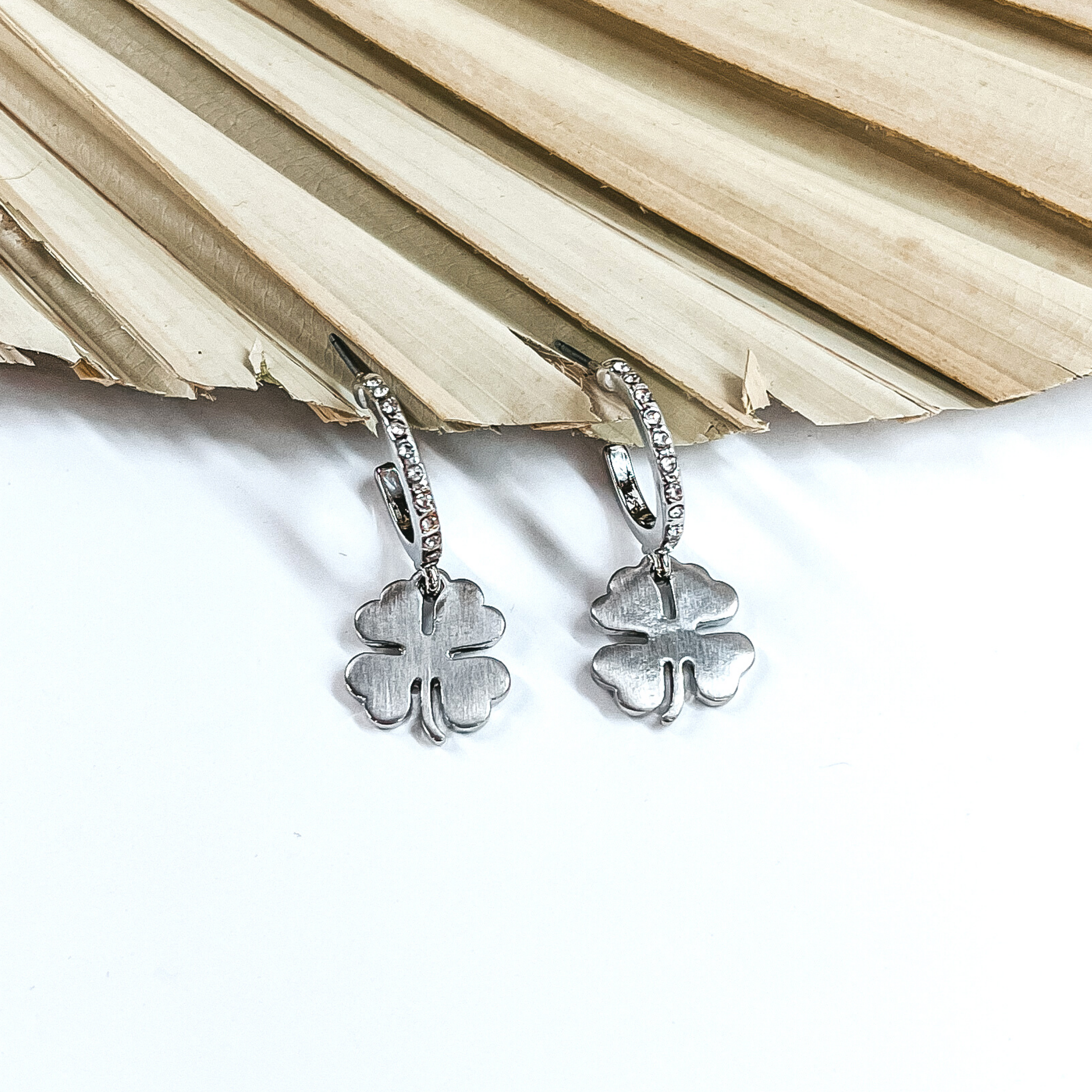 Silver hoops that has a clear crystals. It also has a hanging silver four leaf clover charm. These hoops are pictured in front of a dried leaf on a white background.