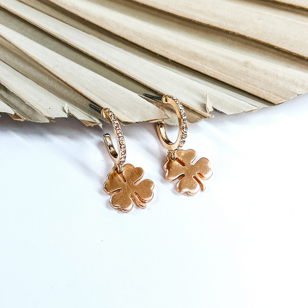 Gold hoops that has a clear crystals. It also has a hanging gold four leaf clover charm. These hoops are pictured in front of a dried leaf on a white background.