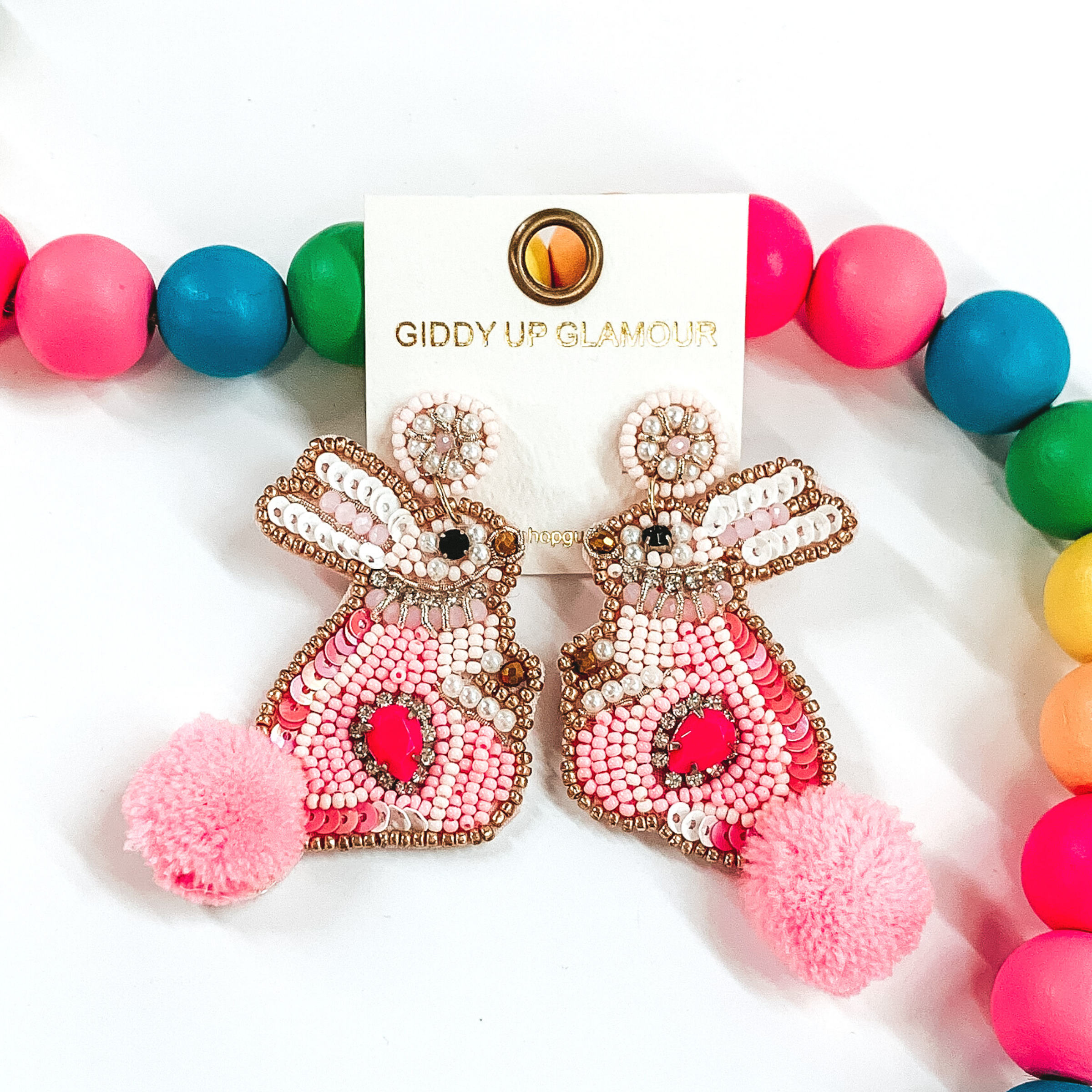 Beaded bunny rabbit shaped earrings. These earrings are outlined in gold beads, with detailing in light and dark pink. The bunny has a pink fluff ball tail. These earrings are pictured in front of some multicolored beads on a white background. 