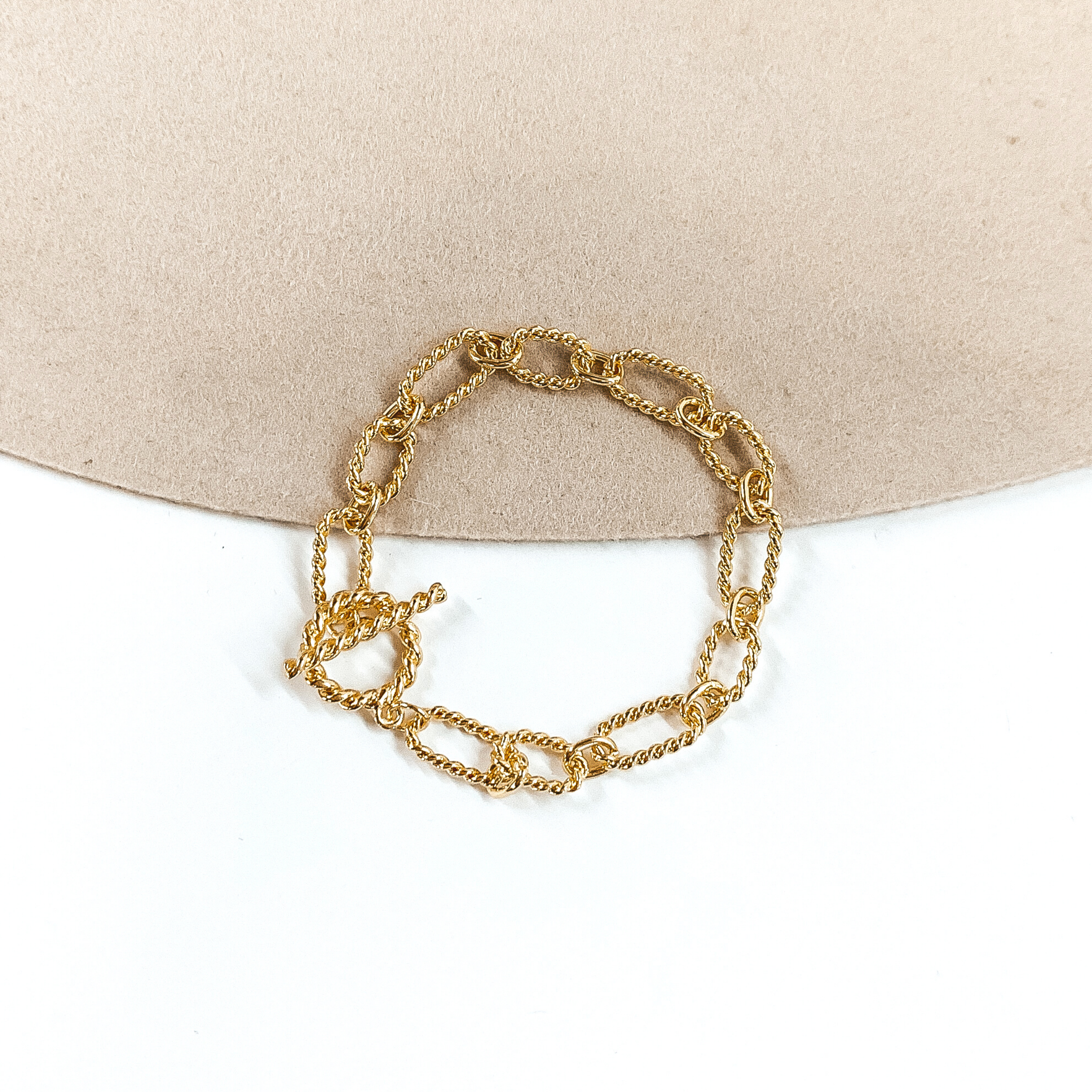 Gold, twisted paperclip chained bracelet in gold. This bracelet has a toggle clasp. This bracelet is pictured on a white and beige background. 