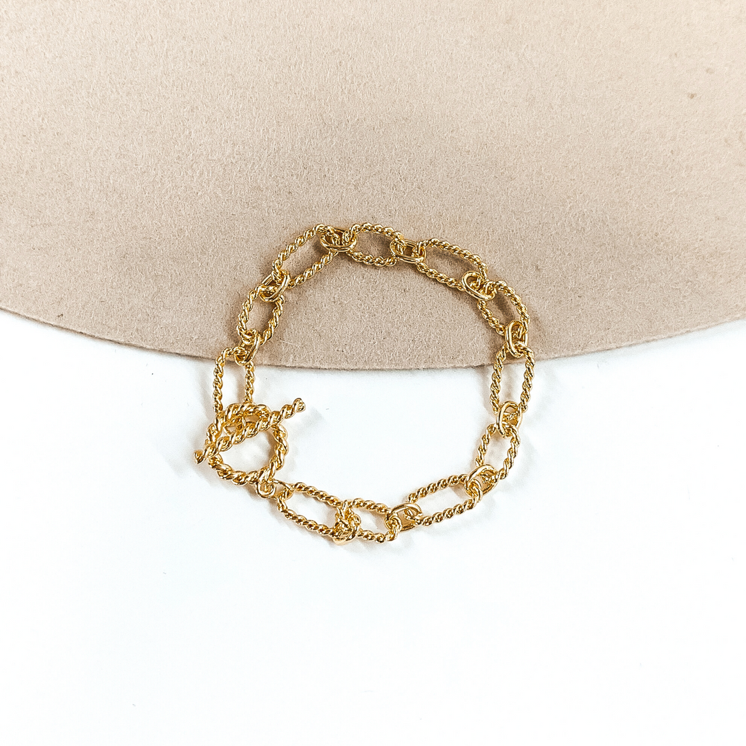 Gold, twisted paperclip chained bracelet in gold. This bracelet has a toggle clasp. This bracelet is pictured on a white and beige background. 